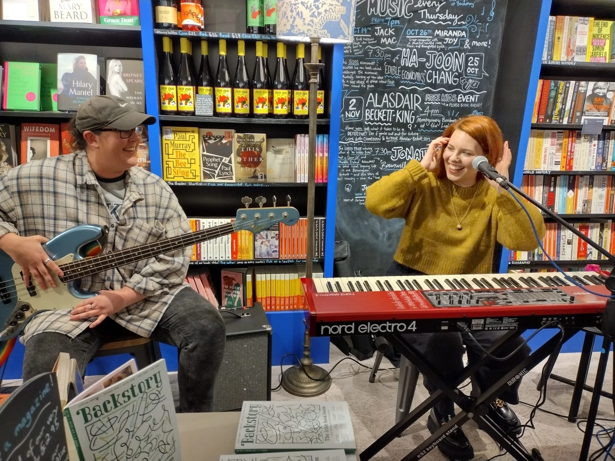 A woman with red hair wearing a yellow sweater is singing into a microphone behind a red Nord Electro 4 keyboard. A man in a checkered shirt, glasses, and cap is sitting to her left, holding a bass guitar and smiling. They are in a bookstore with boo