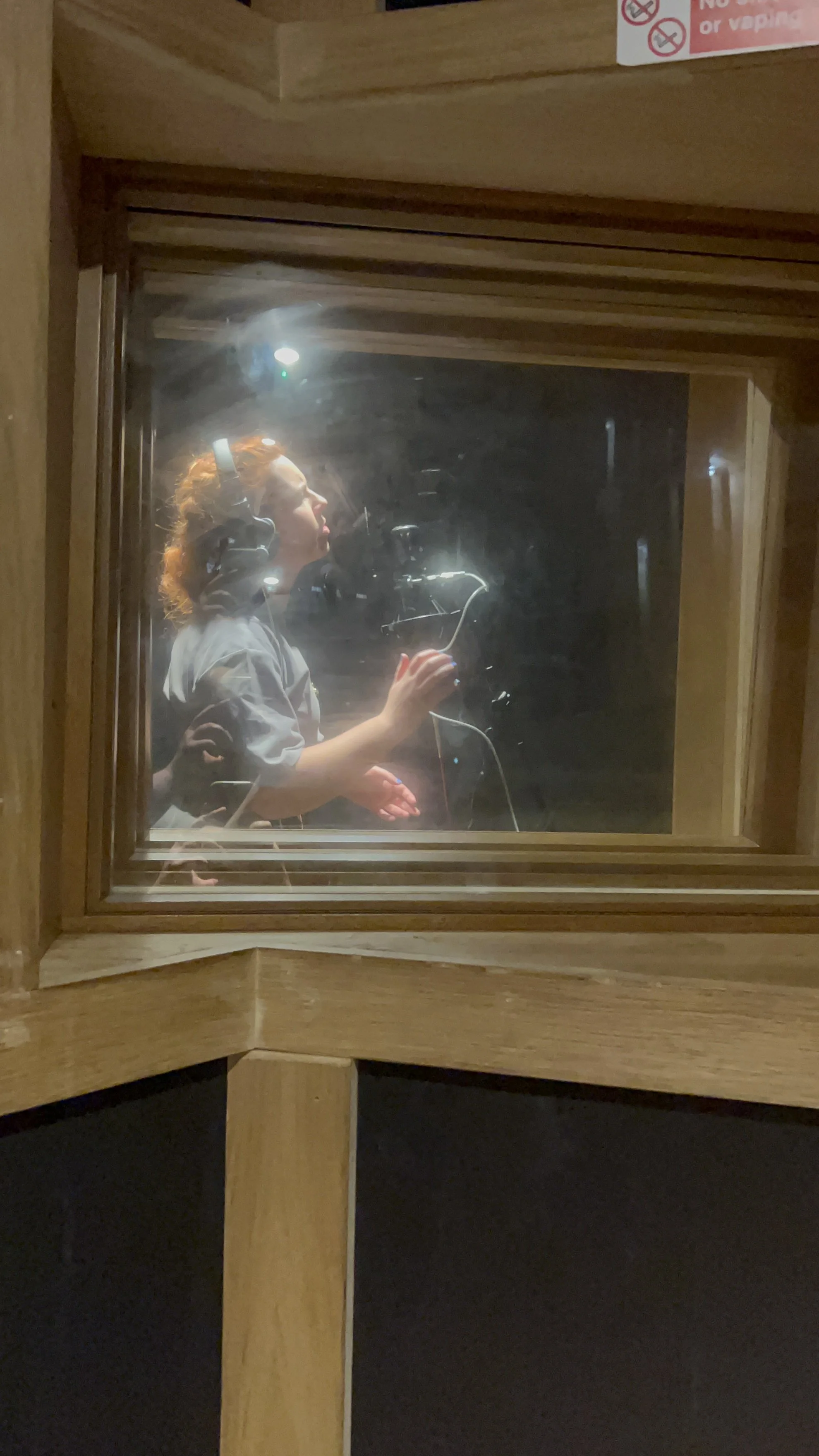 A woman wearing headphones and a gray hoodie is speaking into a microphone in a recording studio, viewed through a window.