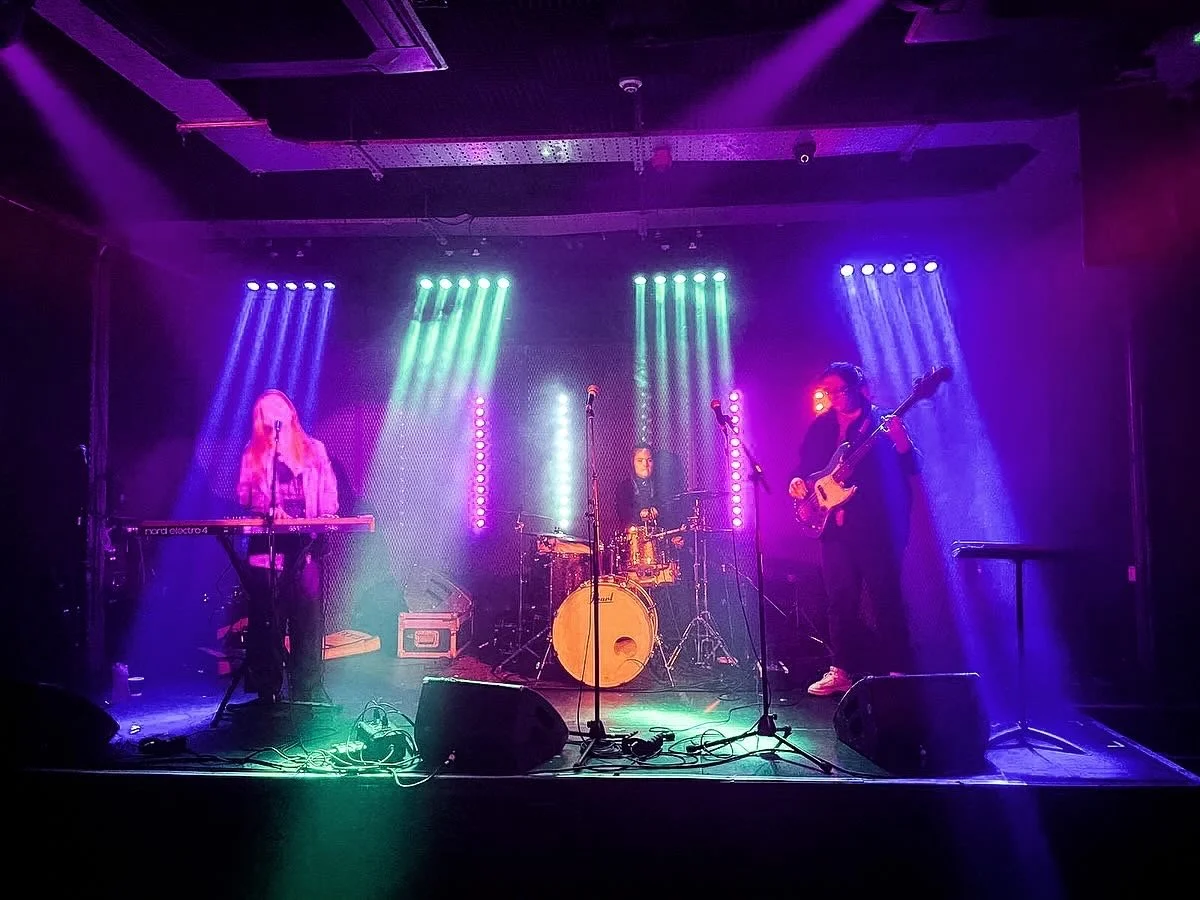 A band of three musicians performing on stage with purple, blue, and green stage lights, including a keyboardist, a drummer, and a bassist.