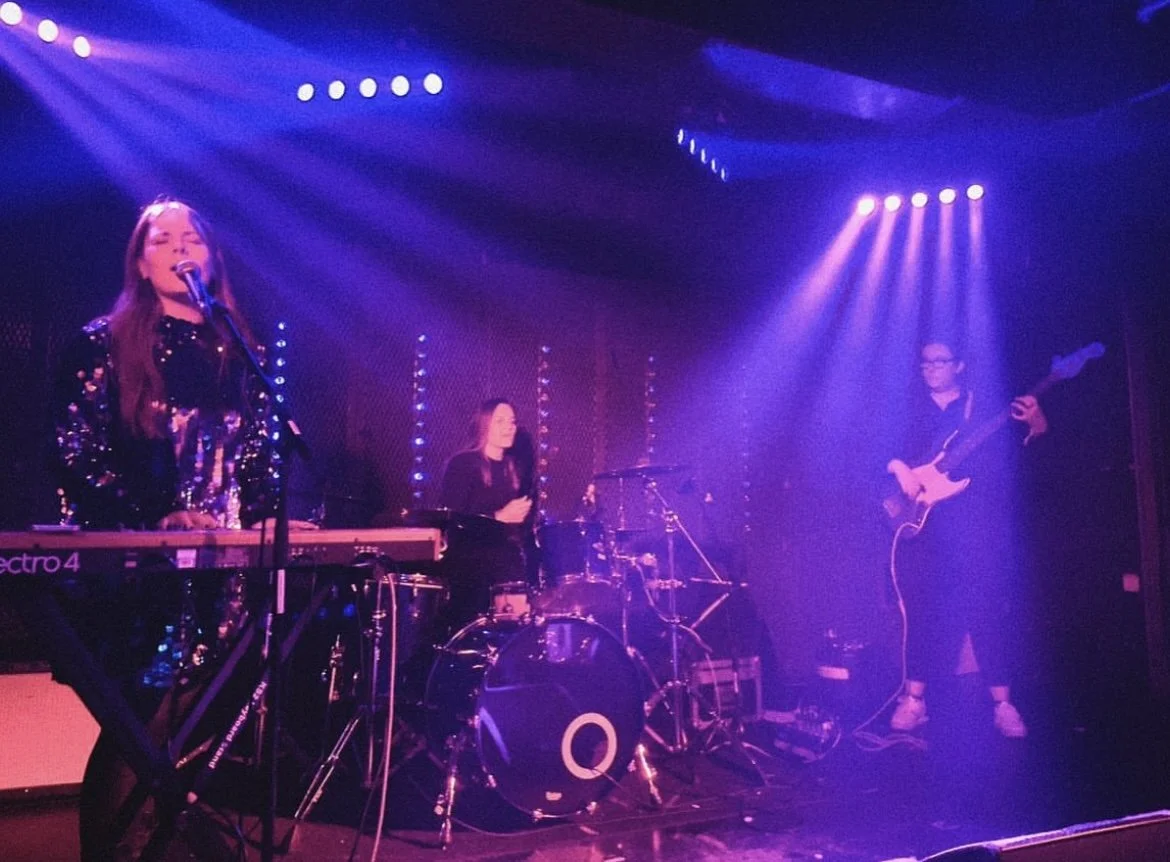 A band performs on stage with purple lighting, including a female keyboardist and singer, a female drummer, and a male guitarist.
