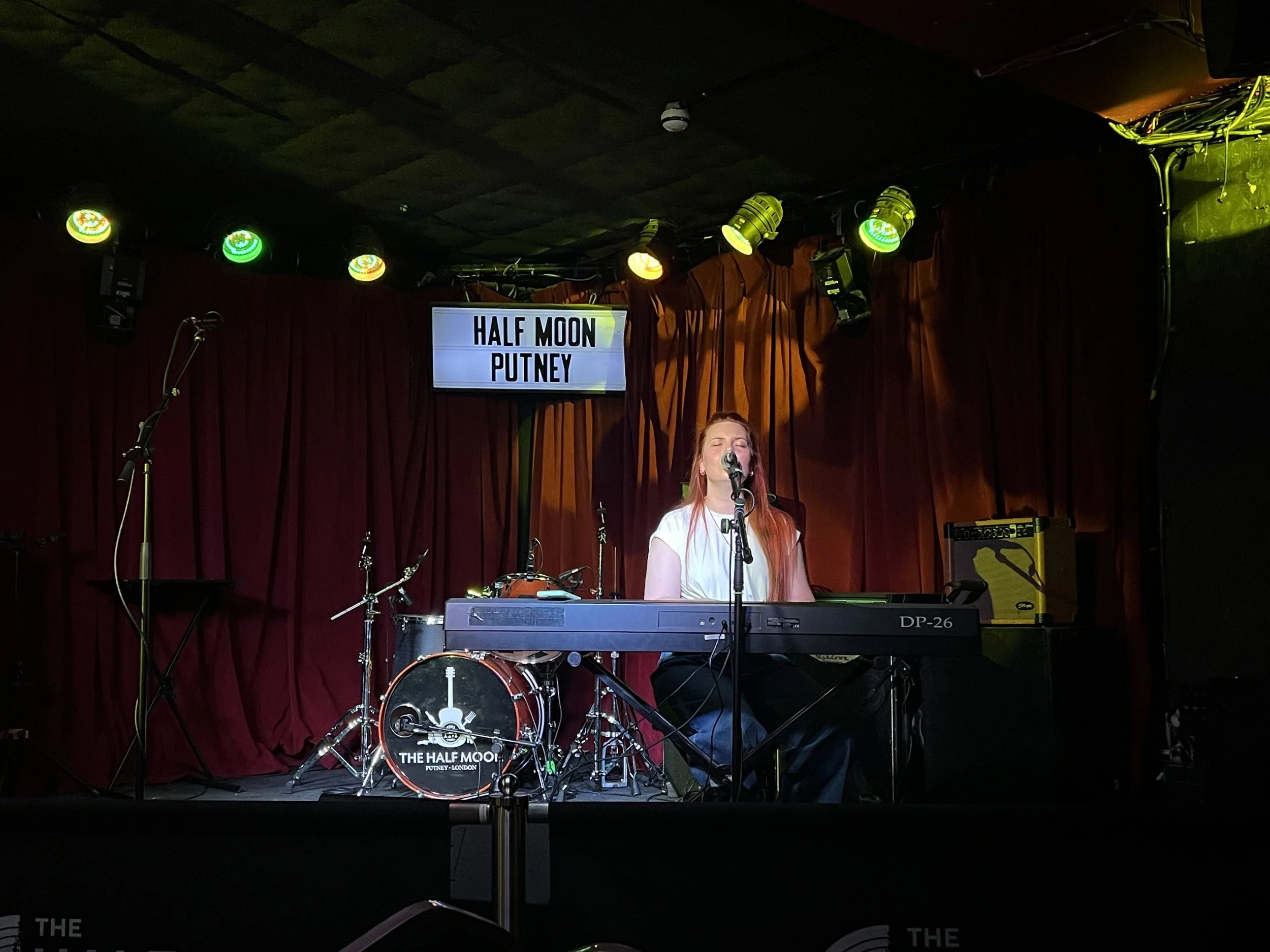 Miranda Joy singing at a keyboard on stage with red curtains and a drum set that has a logo and the words 'The Half Moon Putney London.' A digital sign displays the words 'Half Moon Putney.'
