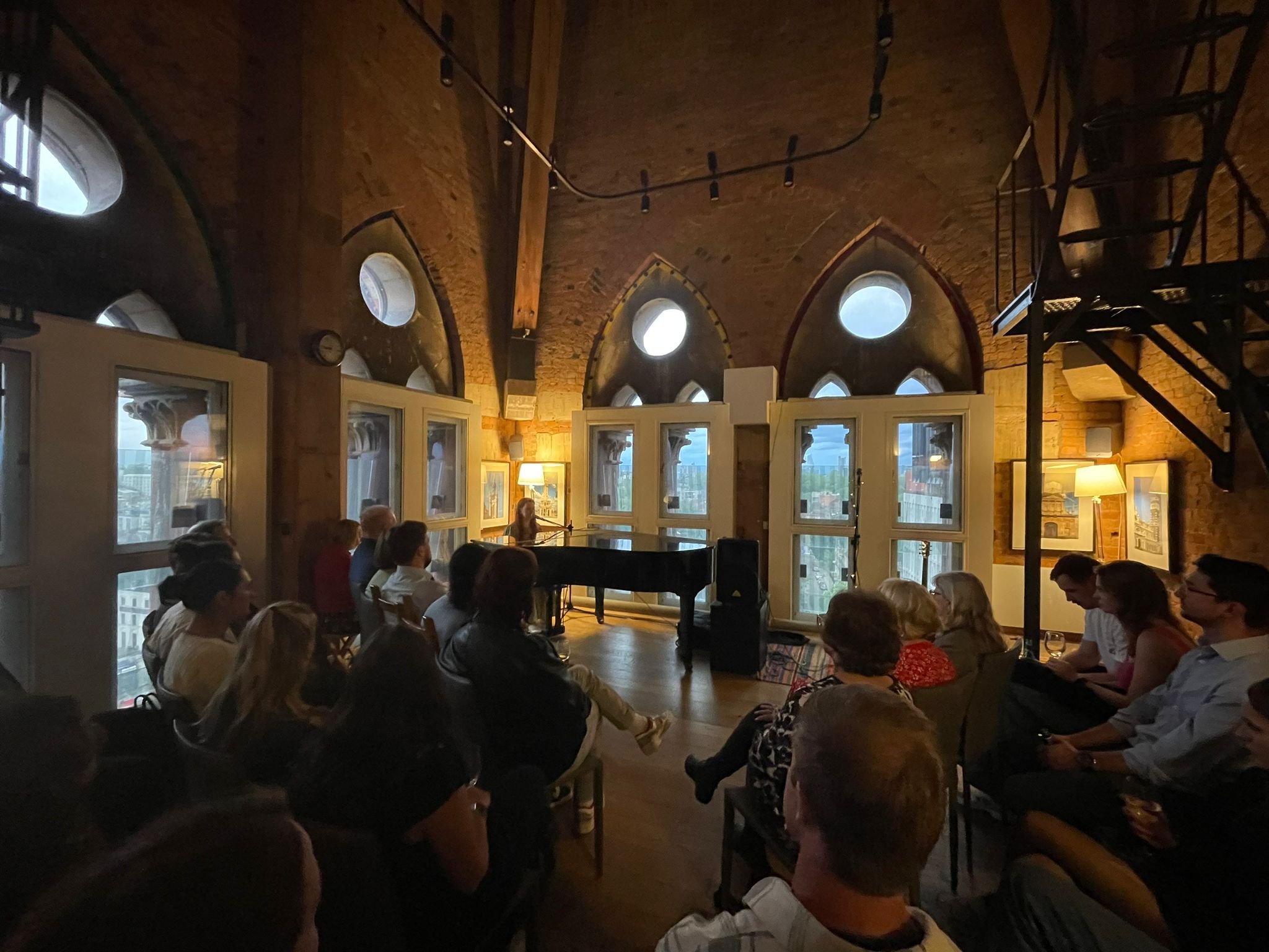A group of people seated in a cozy, brick-walled room with arched windows, watching Miranda Joy playing a piano and singing at a small performance or open mic event.