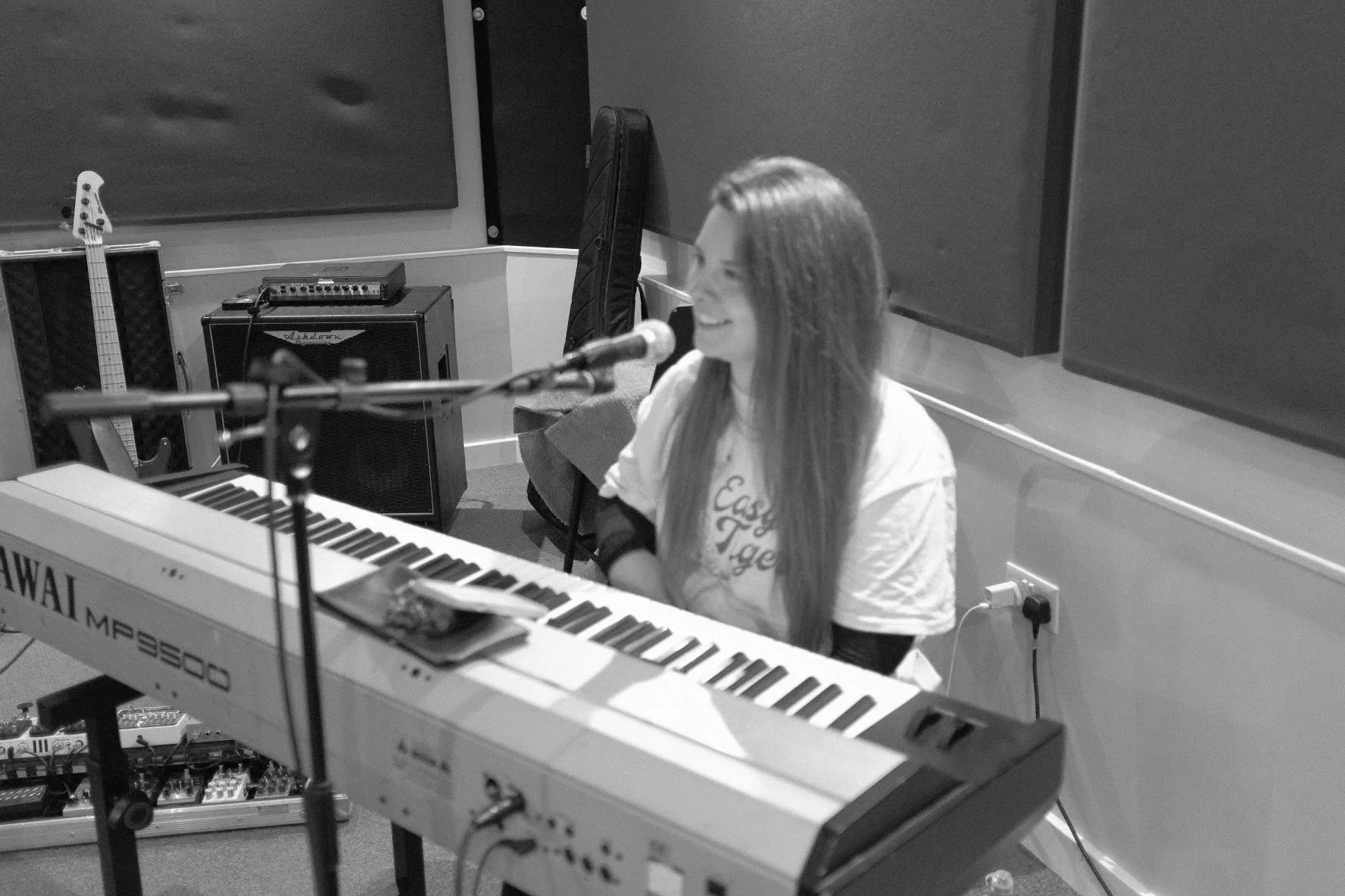 Miranda Joy sitting at a keyboard in a music studio, smiling, with microphone in front of her, and musical equipment around.
