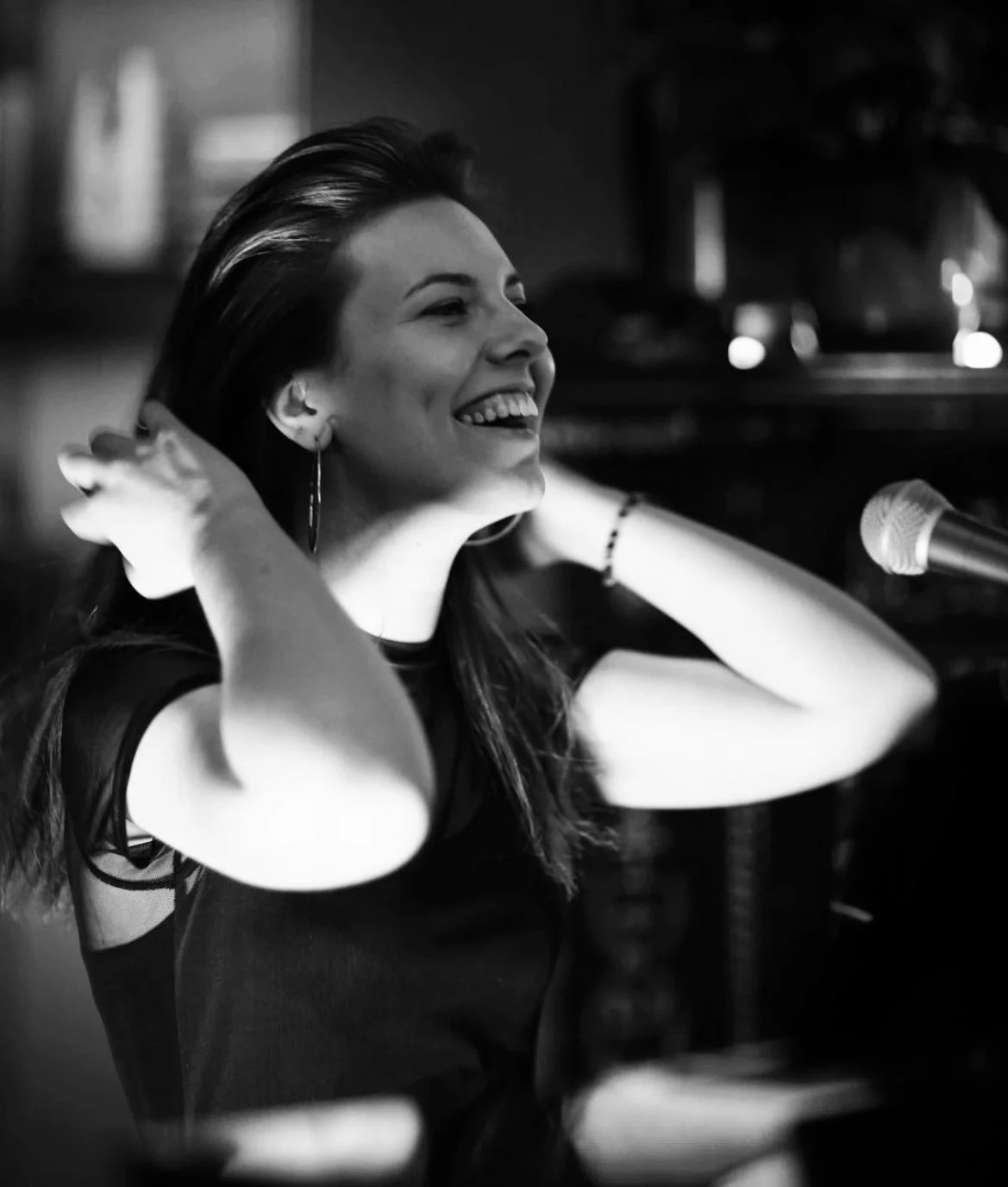 A woman with dark hair smiling and touching her hair, sitting in front of a microphone in a dimly lit environment, captured in black and white.