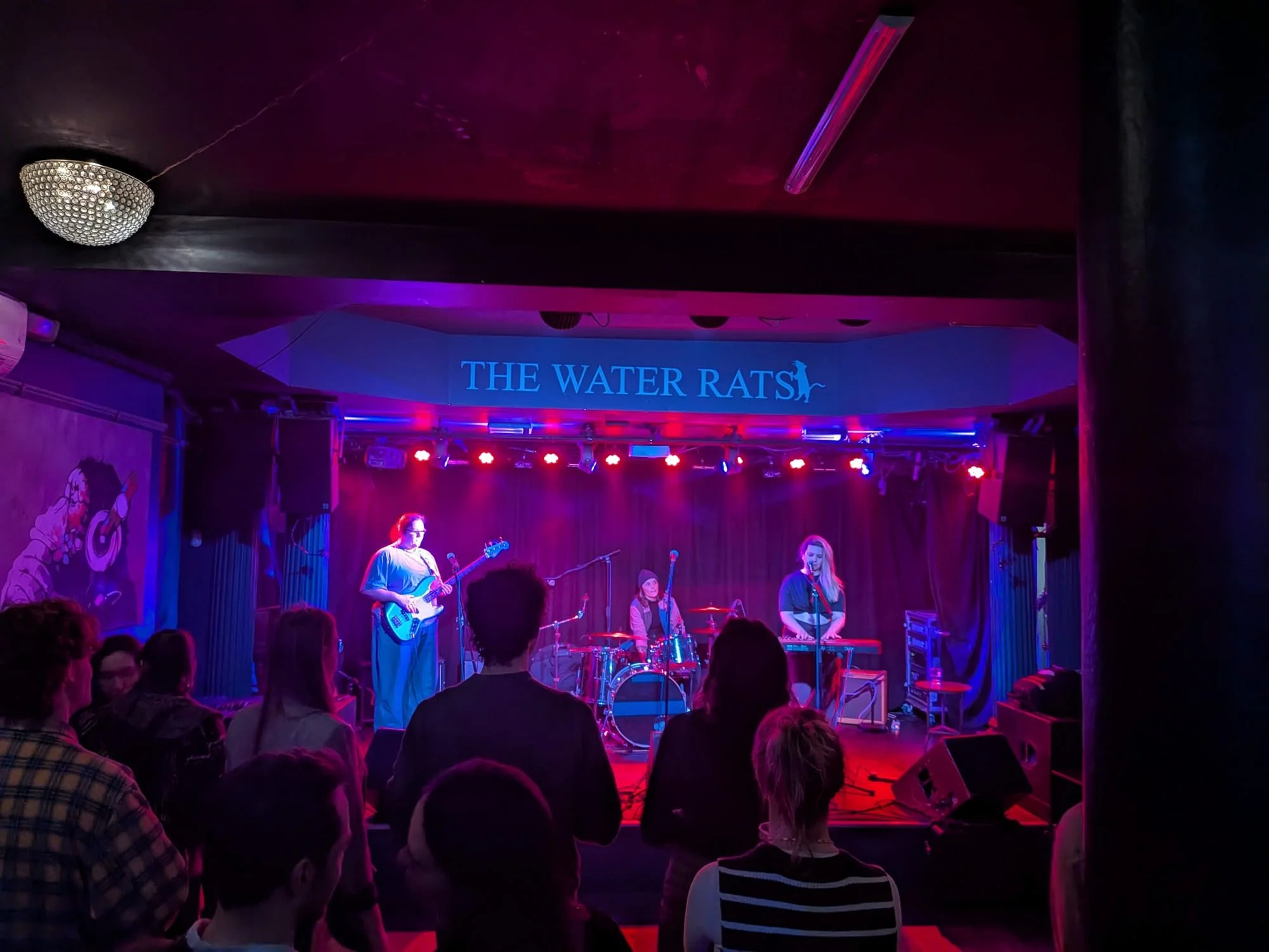 A live musical performance on stage at The Water Rats, with Miranda Joy and her band members playing instruments and singing, audience members watching in front, dim lighting with pink and blue neon lights.
