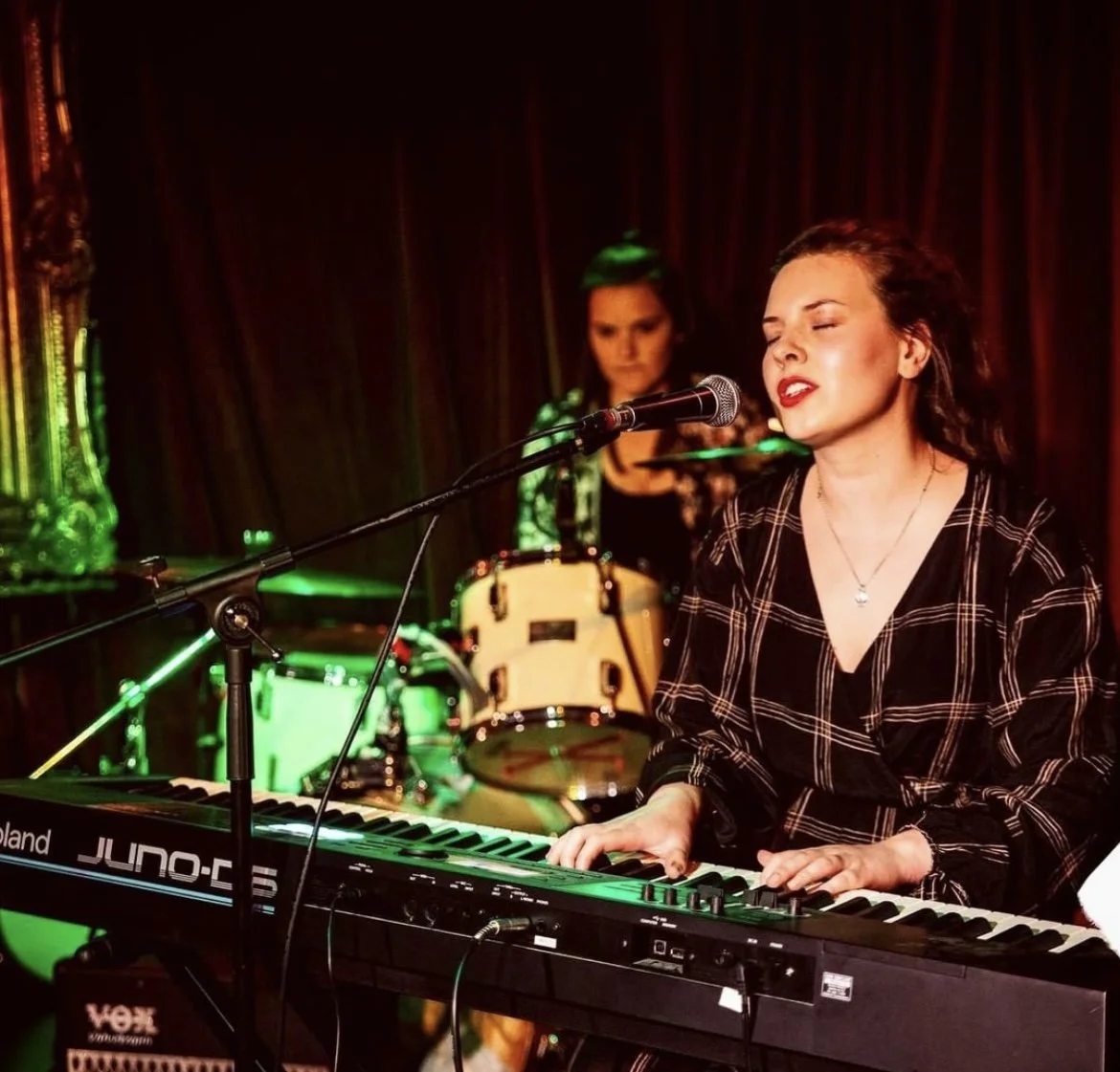 A woman singing and playing a keyboard on stage with a drummer in the background, performing in a dimly lit venue with dark curtains and colorful stage lights.