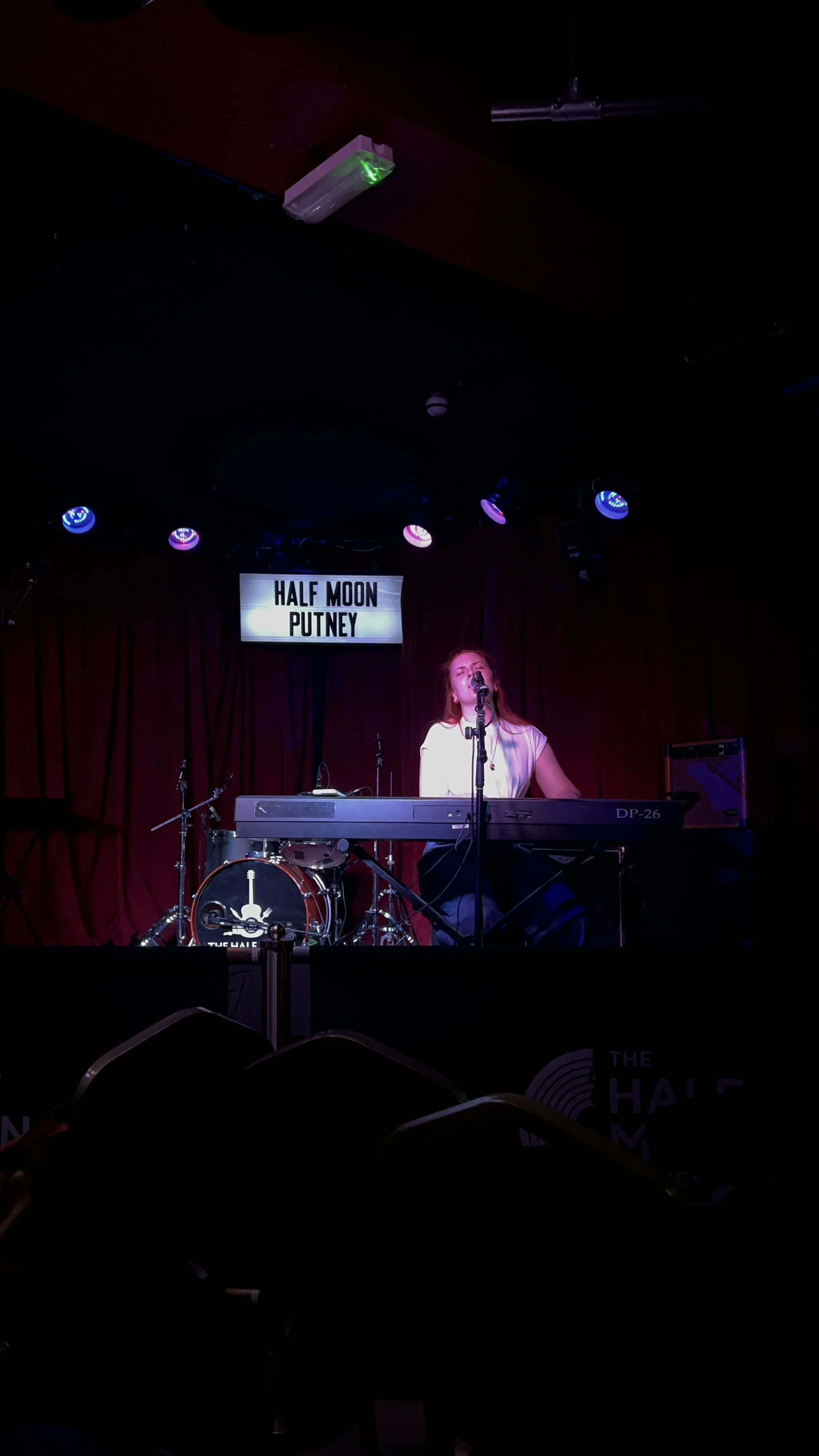 A woman performing on stage at Half Moon Putney, sitting at a keyboard with a microphone, with a drum set in the background, red curtains, and stage lights overhead.