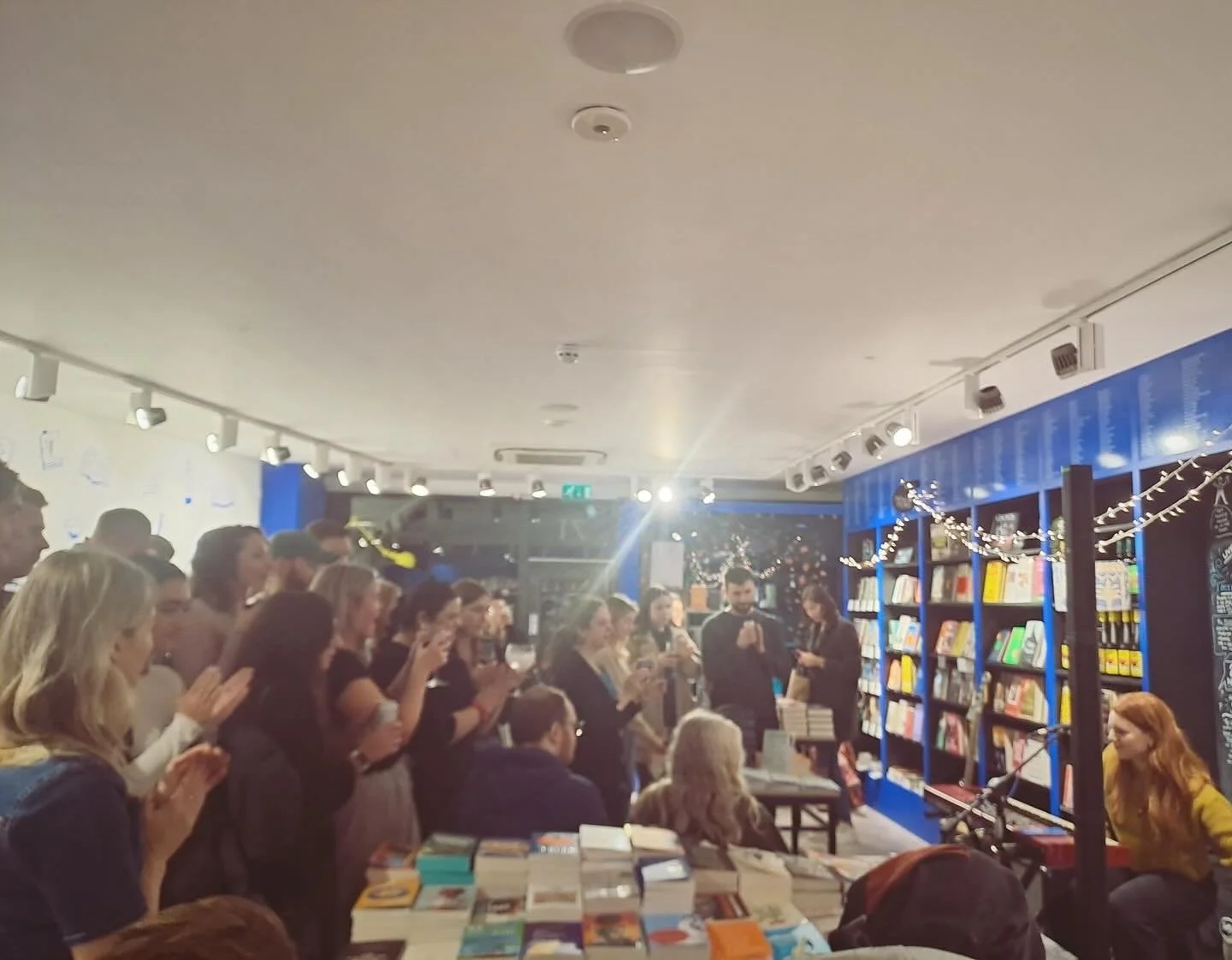 People gathered in a bookstore or event space, some clapping and others using their phones, with shelves of books and holiday string lights in the background.
