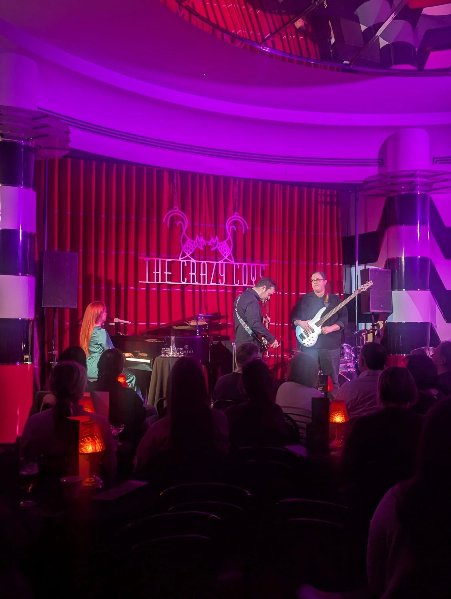 Miranda Joy and band performing on stage at The Crazy Cock, bright purple and red lighting, audience seated with glowing table lanterns.