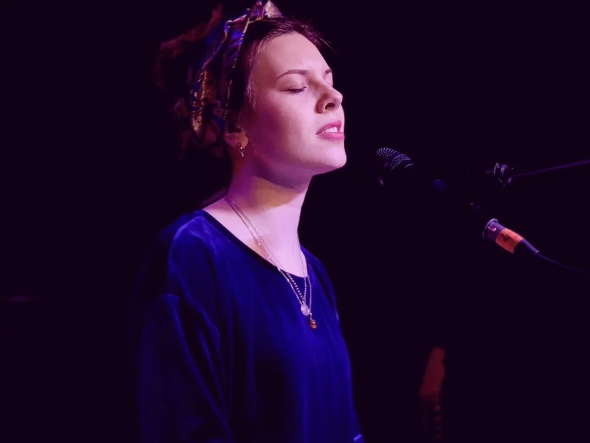 A woman with braided hair singing into a microphone with closed eyes in a dark setting illuminated by purple light.