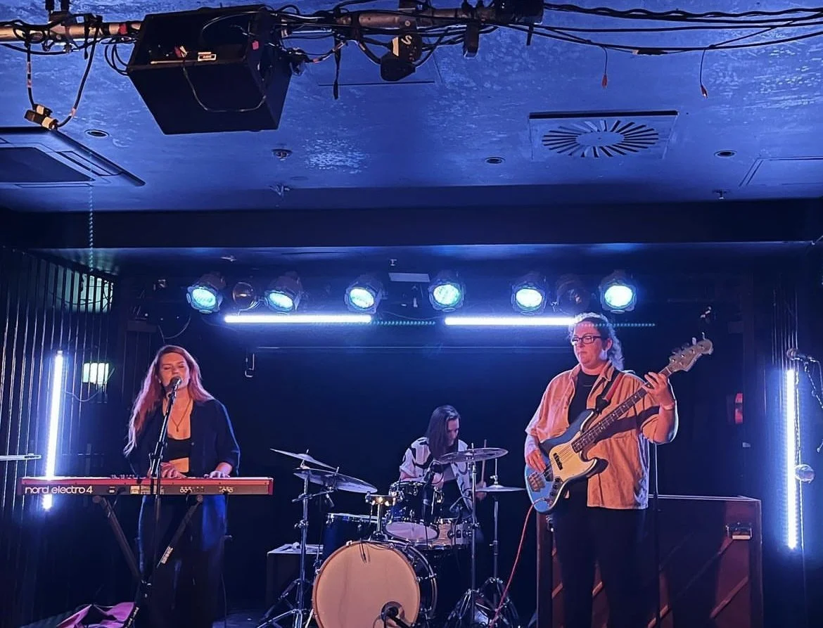 Three women perform on a stage, one playing a keyboard, another singing, and the third playing a bass guitar. The stage is lit with blue and white lights, and musical equipment is visible.