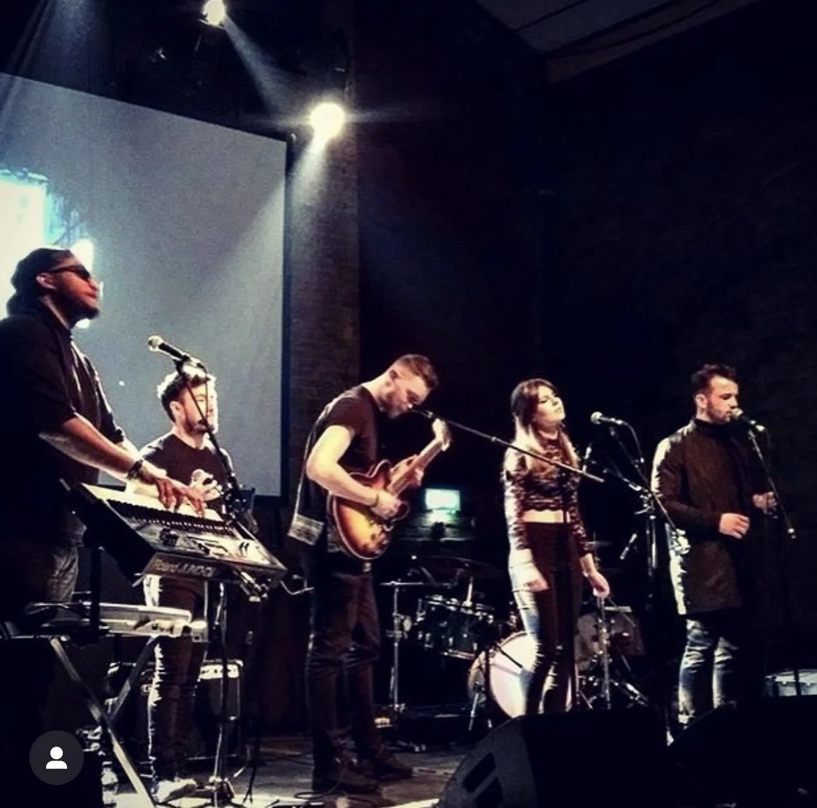 A band performing on stage with five musicians playing various instruments and singing into microphones, in a dimly lit venue.