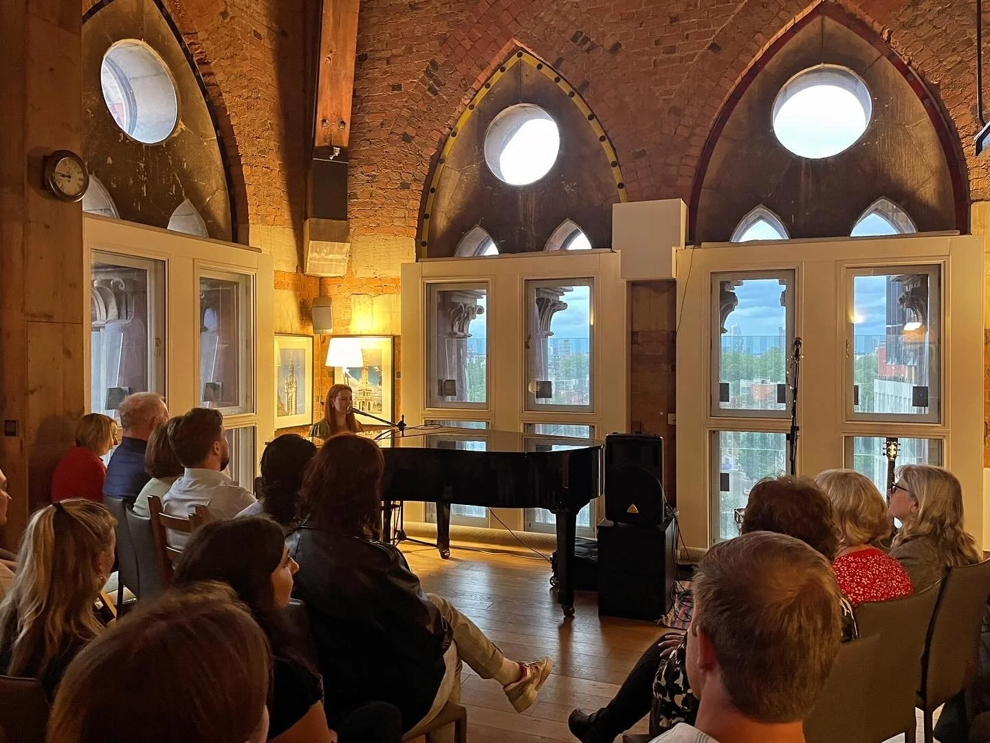 Miranda Joy is performing on a black grand piano in a rustic, brick-walled room with large windows and circular windows high above. An audience is seated facing her, listening attentively. There is a lamp on the wall and framed artwork.