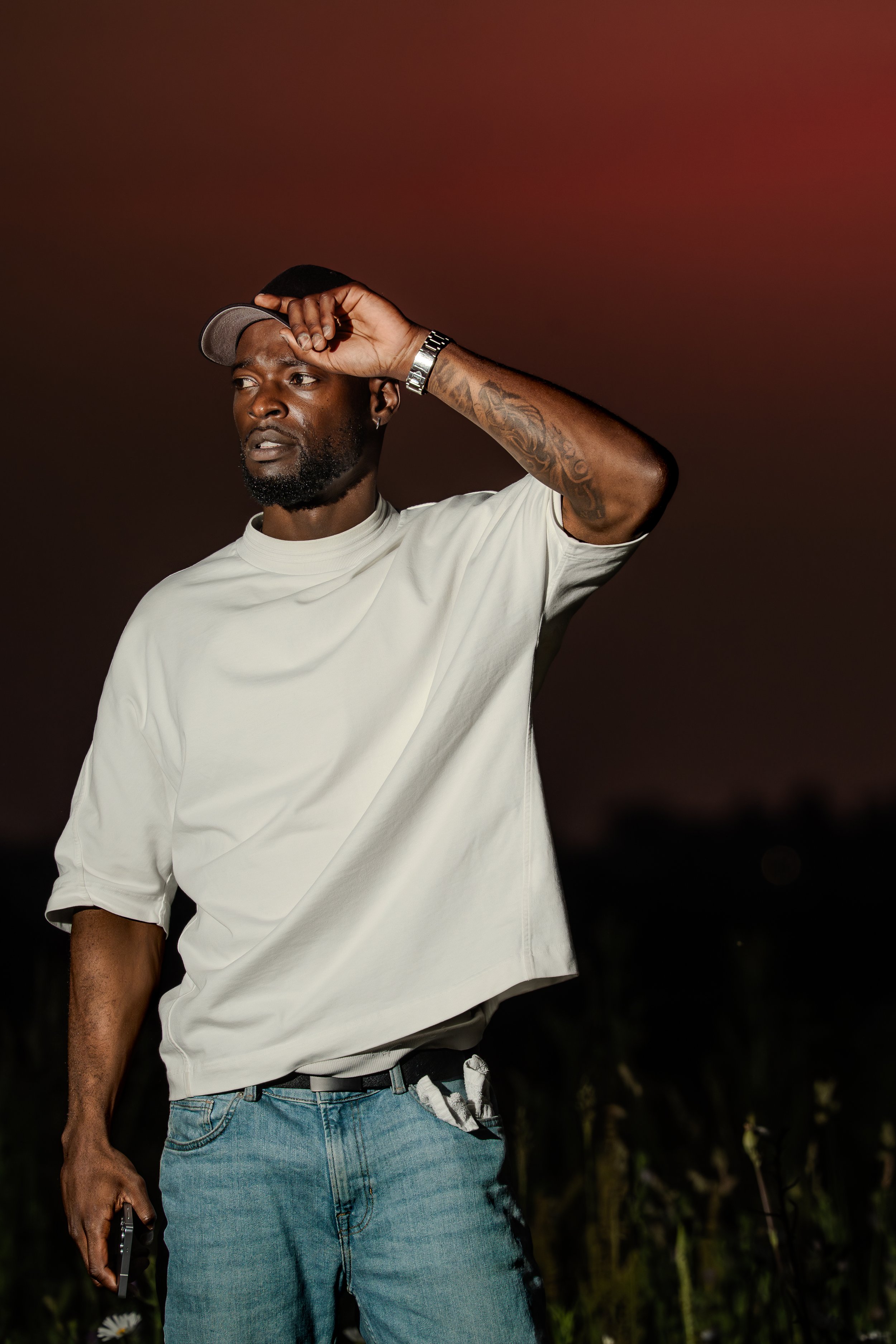 Homme noir portant un t-shirt blanc, un jean bleu et une casquette, tenant un téléphone mobile, avec un tatouage sur le bras, le tout dans un environnement extérieur au crépuscule.