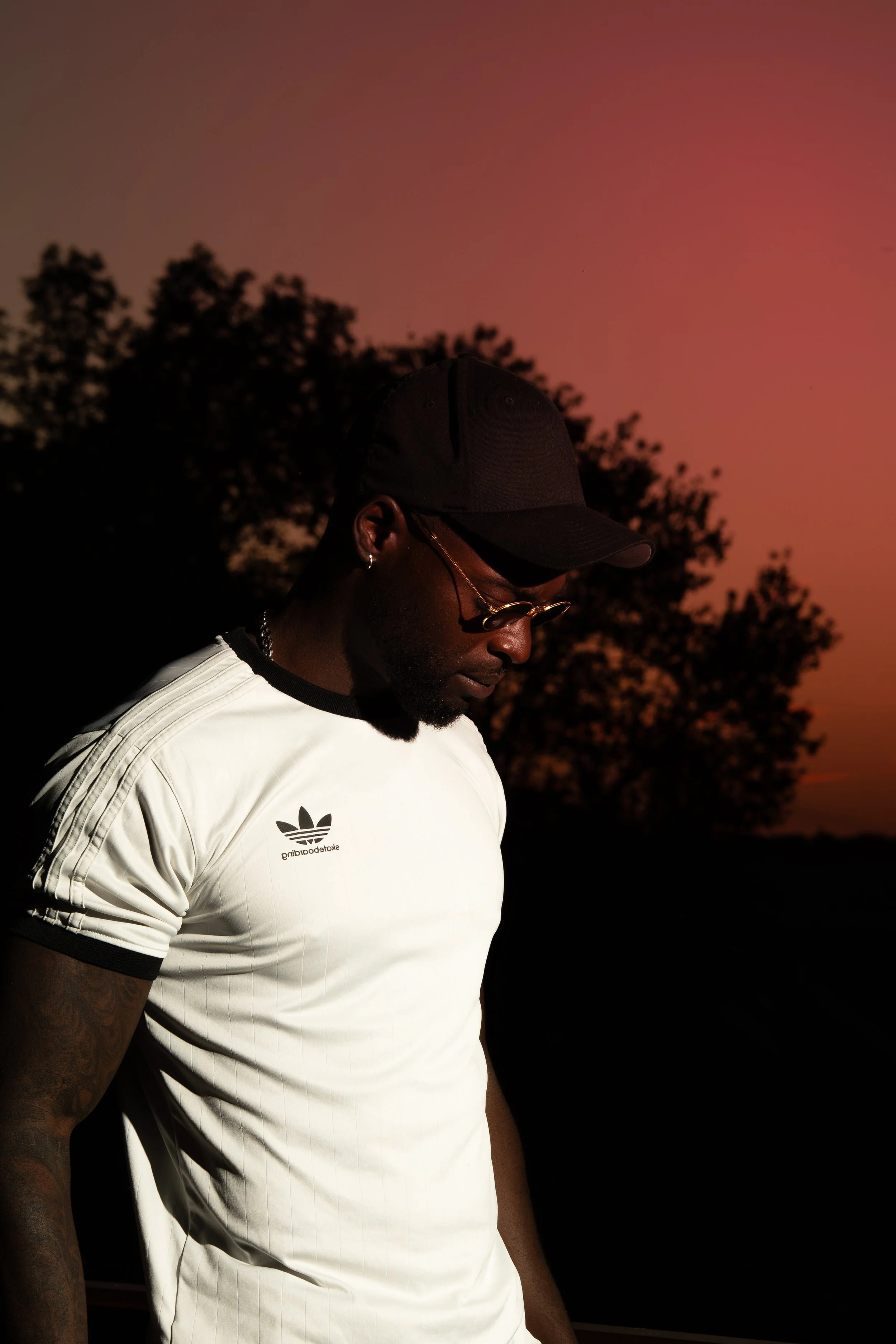 Homme portant une casquette noire, des lunettes de soleil, une chaîne, un petit anneau à l'oreille, et un t-shirt blanc avec le logo Adidas, regardant vers le bas lors d'un coucher de soleil avec un ciel rose et orange, silhouette d'arbres en arrière-plan.