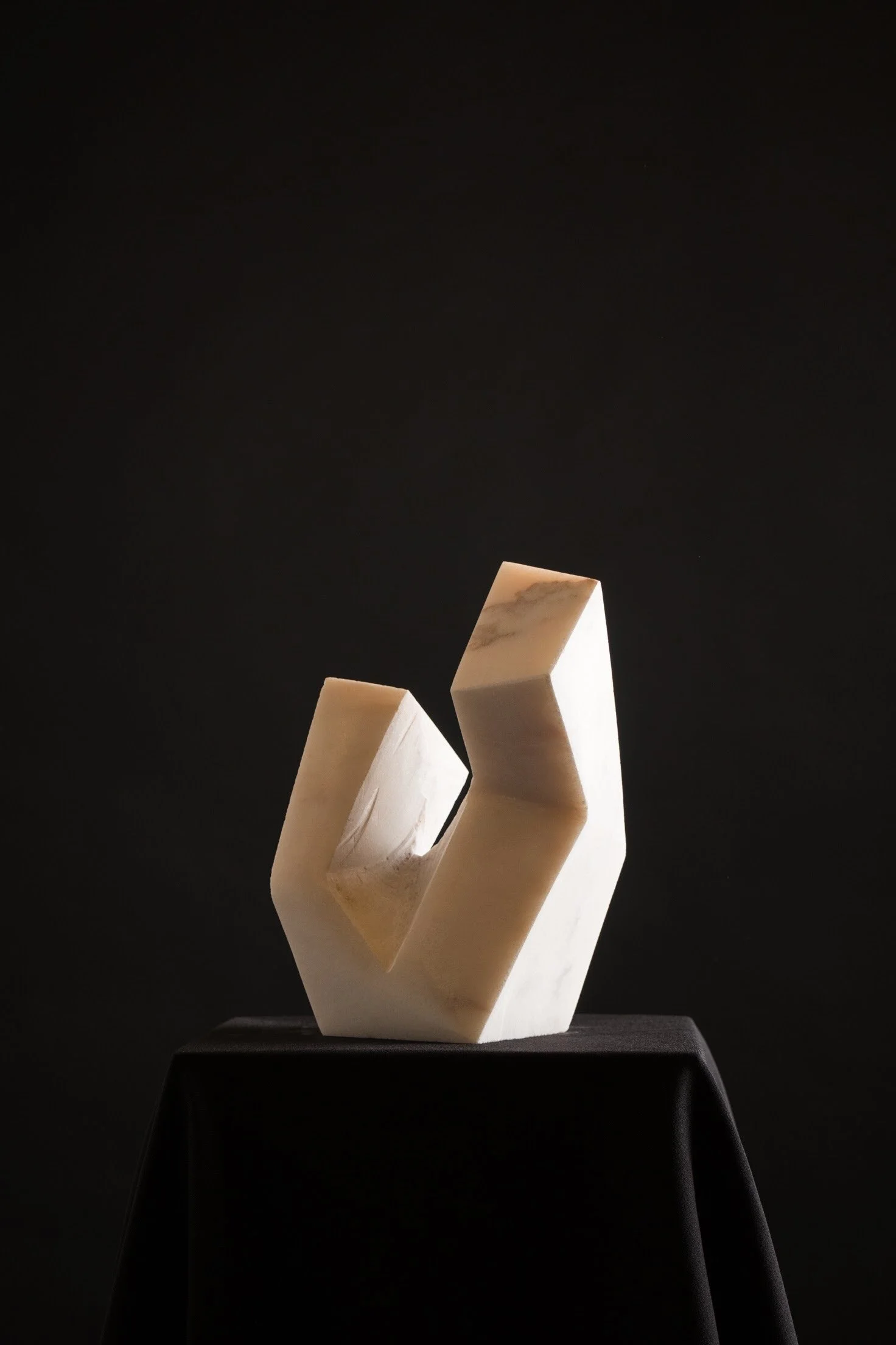 Abstract white sculpture with geometric shapes on a black pedestal against a dark background.