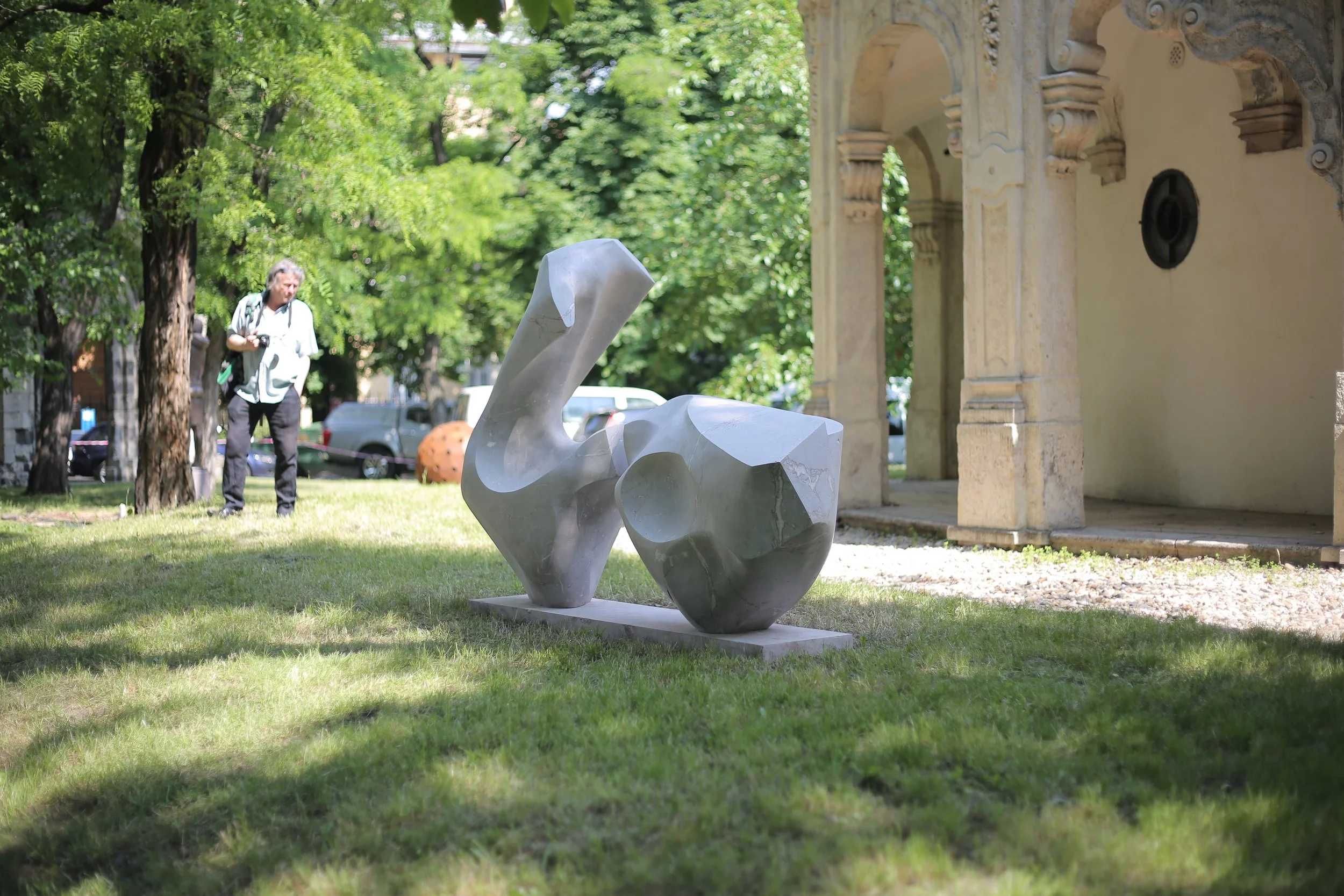 Contemporary abstract sculpture made of smooth gray stone on a grassy area, with trees and a building in the background, and a person walking with a camera in the distance.