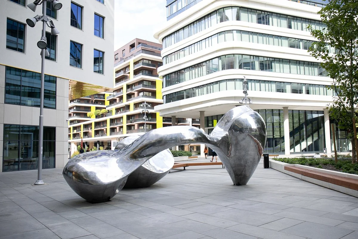 Modern outdoor sculpture resembling a large, abstract, metallic form in an urban plaza with buildings and trees in the background.