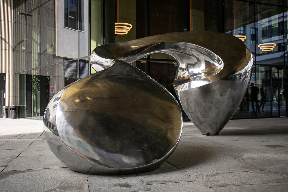 A modern abstract sculpture made of shiny metal, featuring smooth, curved shapes, displayed outside a building with glass windows and modern lighting fixtures.