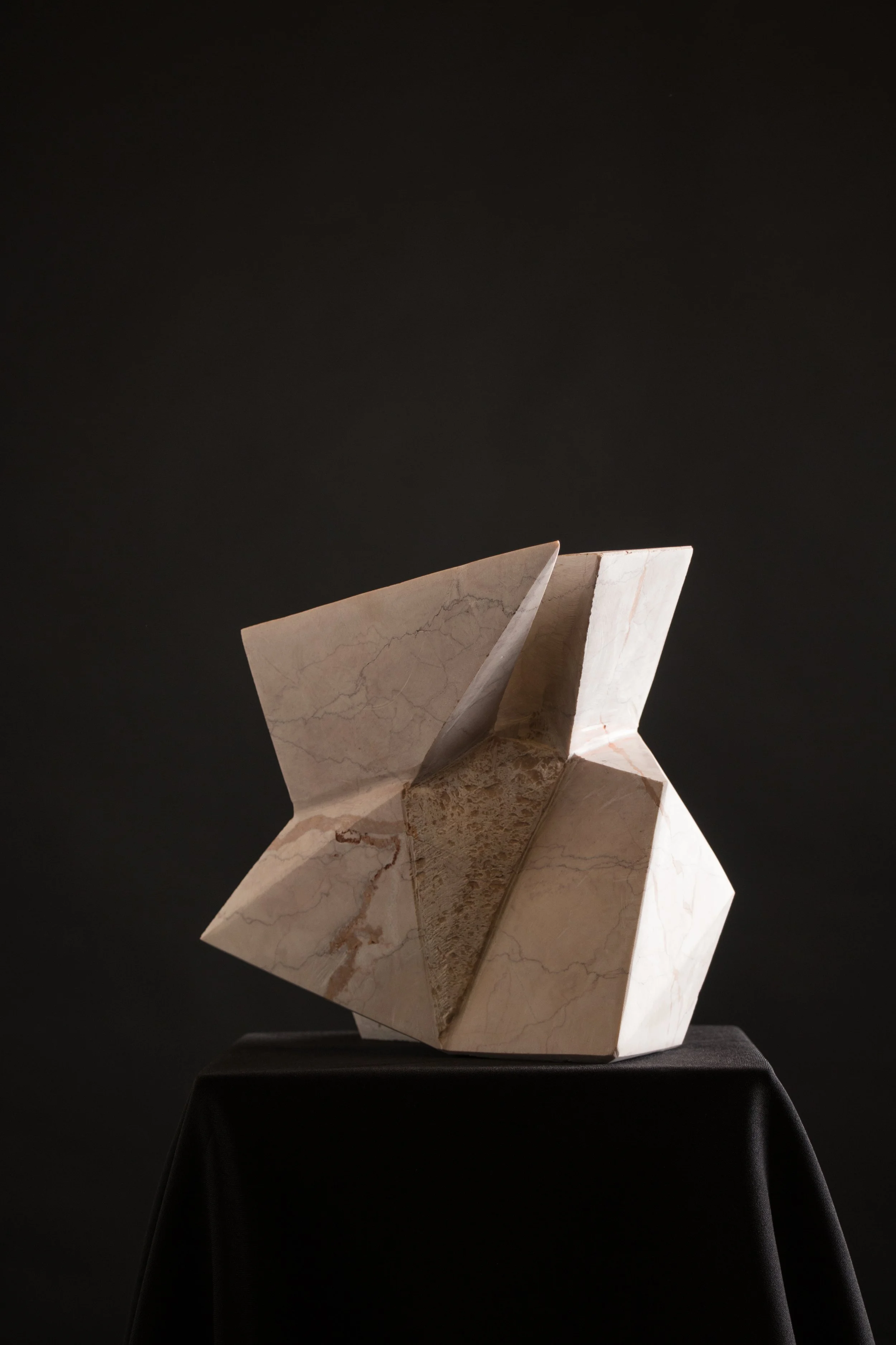 Abstract marble sculpture with angular geometric shapes on a black cloth-covered pedestal against a dark background.
