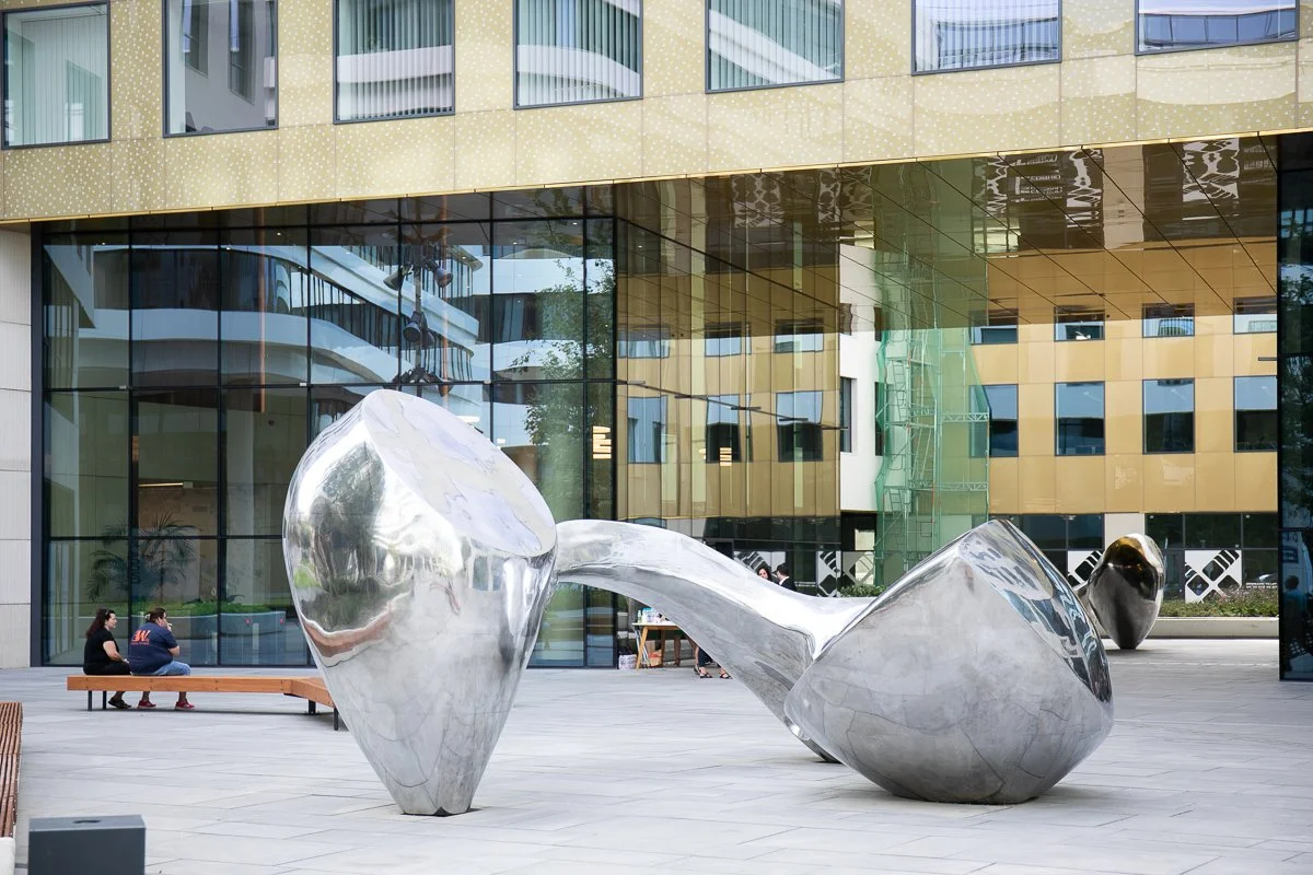 Large reflective abstract sculpture made of shiny metal in an urban plaza with modern glass and gold-colored building in the background, two people sitting on a bench nearby.