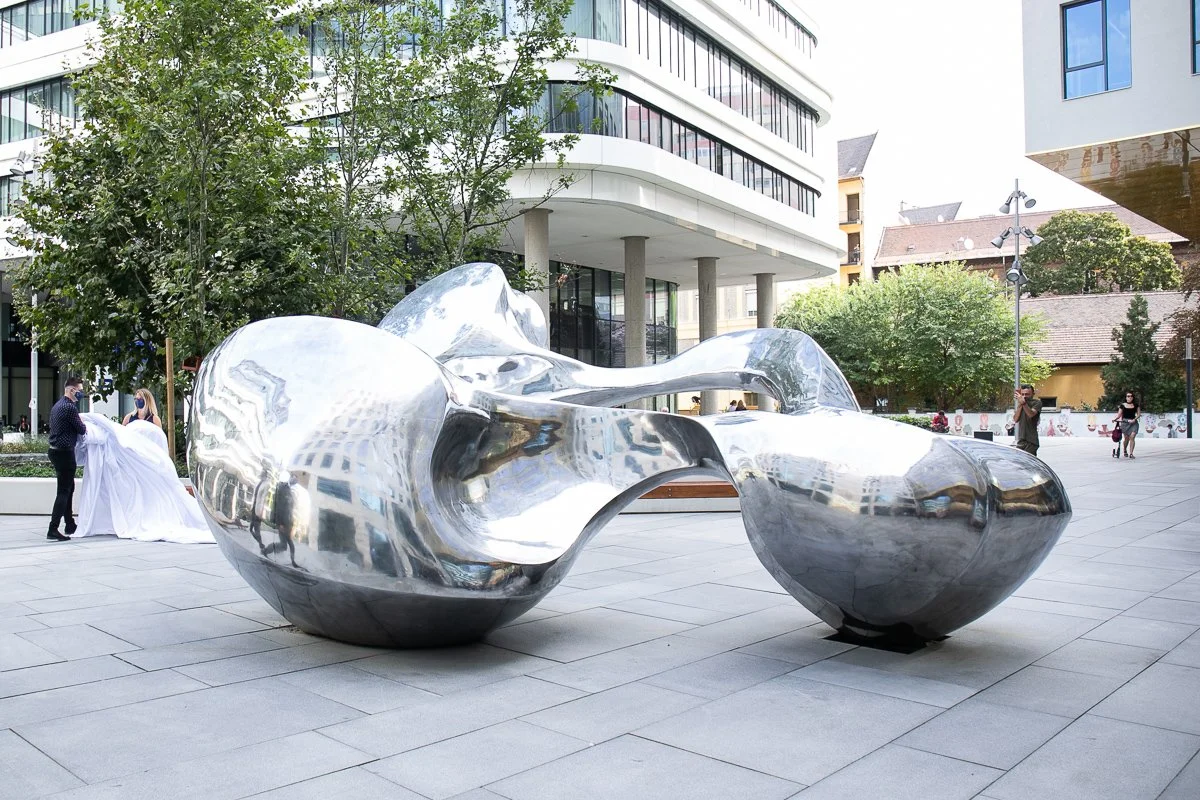 Large shiny metal abstract sculpture in an urban plaza with modern buildings, trees, and people nearby.