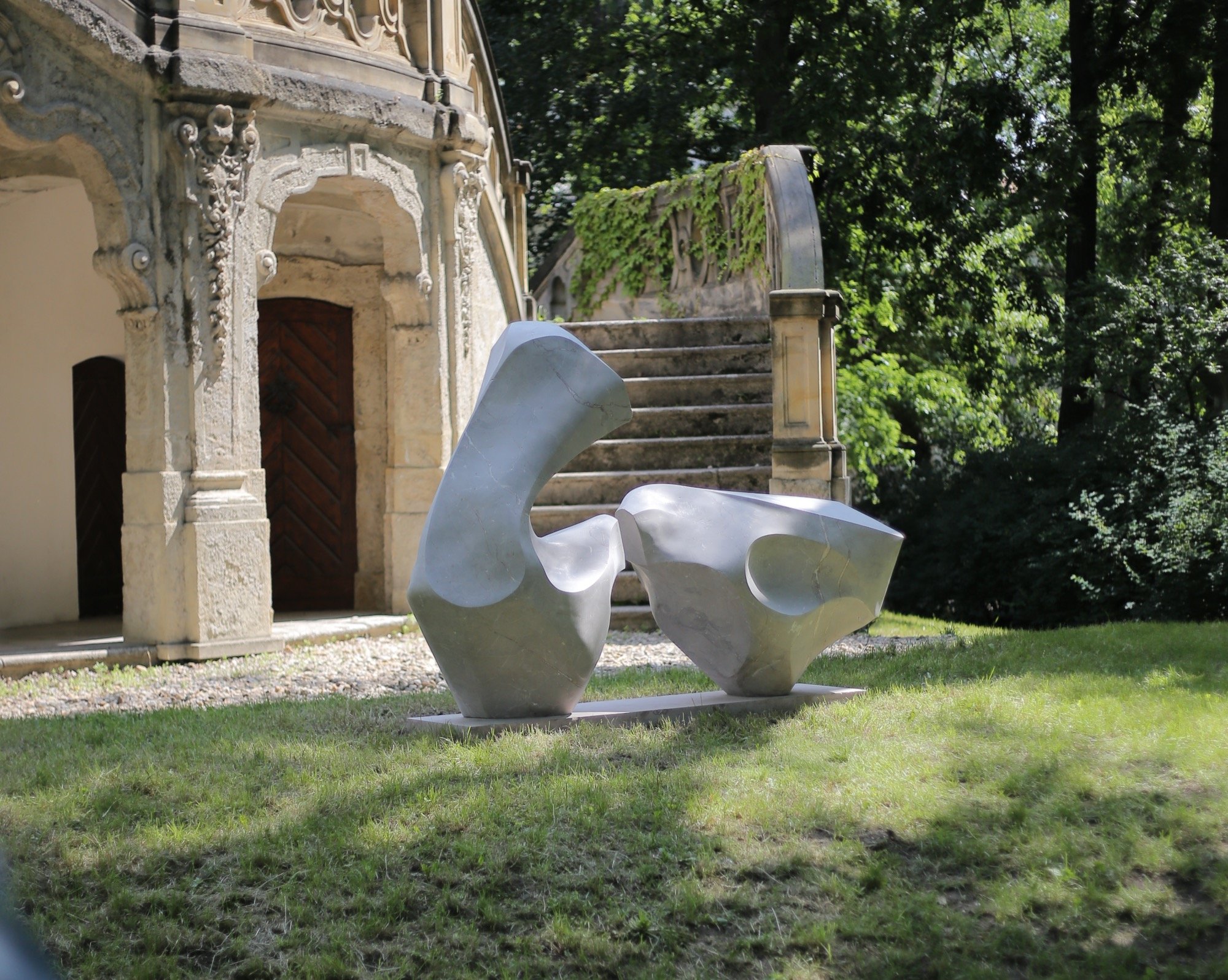 An abstract metallic sculpture with smooth, curving surfaces situated on a grassy area in front of an ornate stone building with stairs and lush green trees in the background.