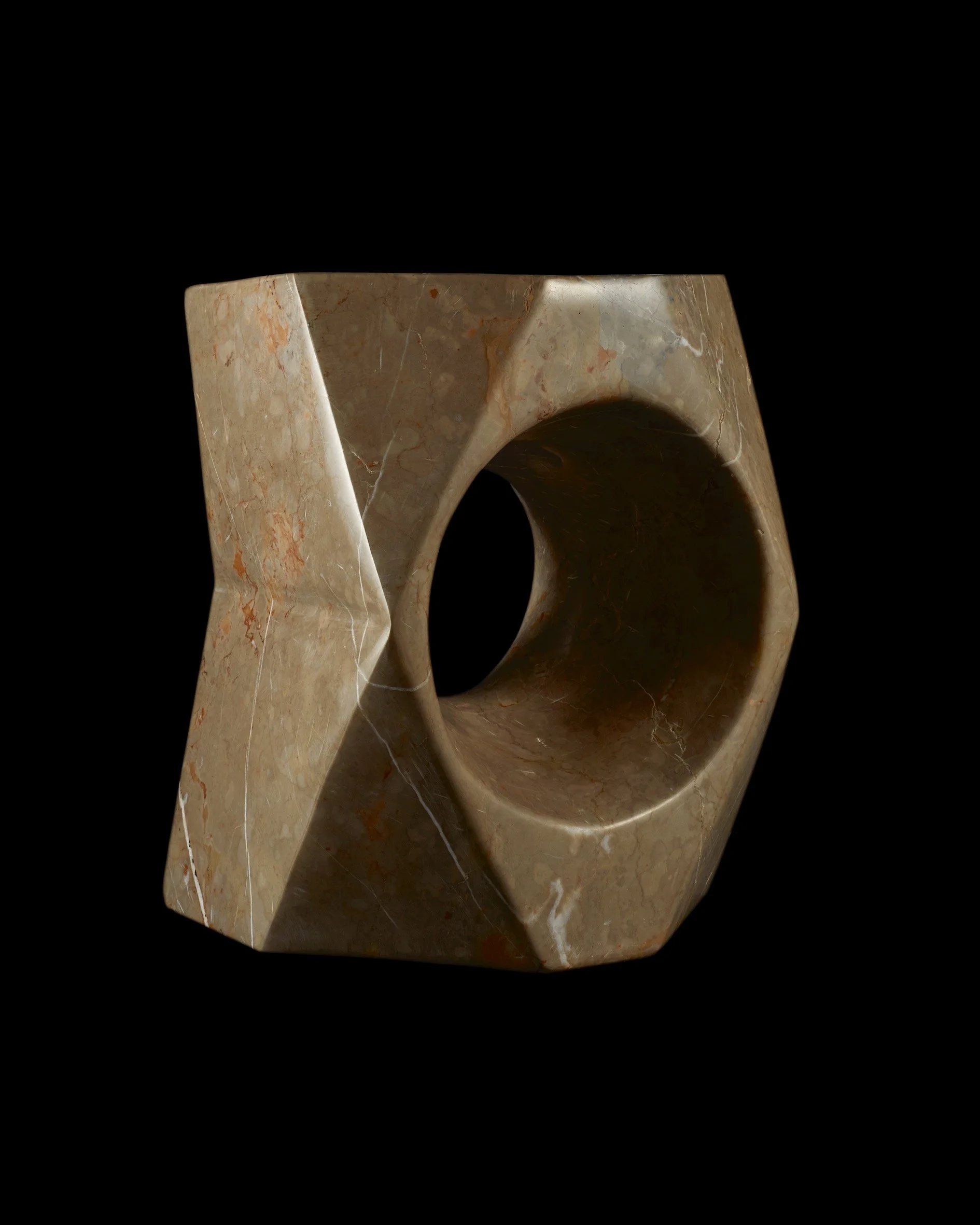 A large, irregularly shaped, beige and brown marble sculpture with a circular hole in the center, set against a black background.