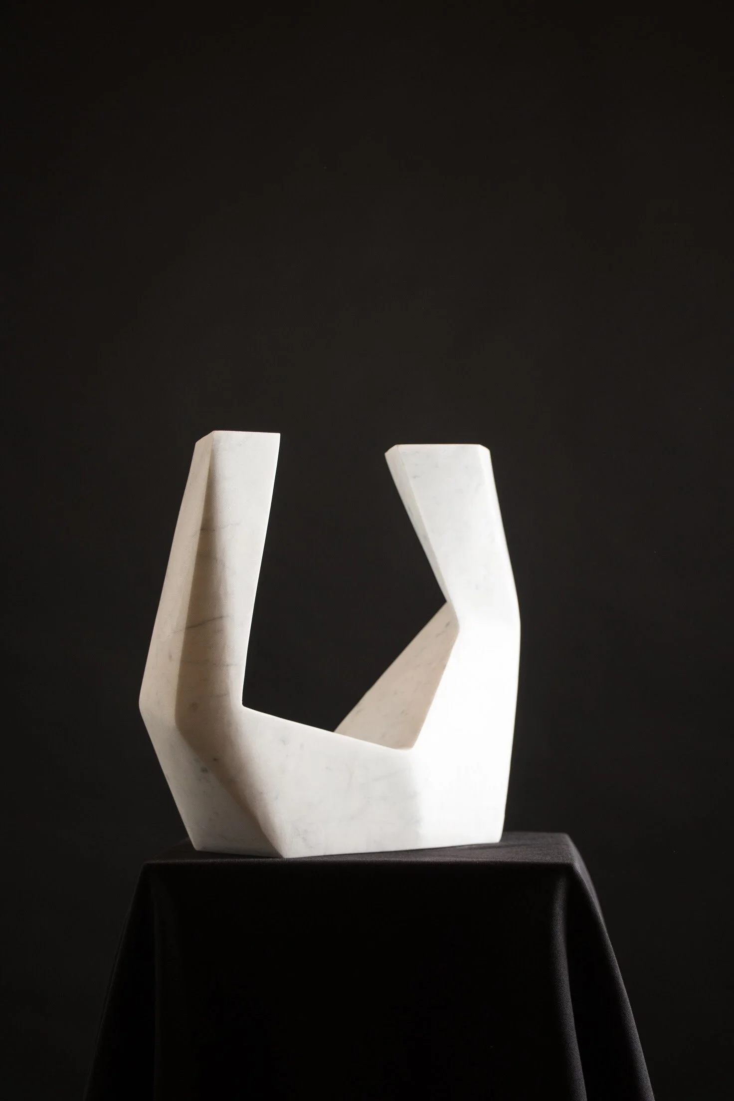 A modern white marble sculpture with geometric shapes, displayed on a black pedestal against a black background.