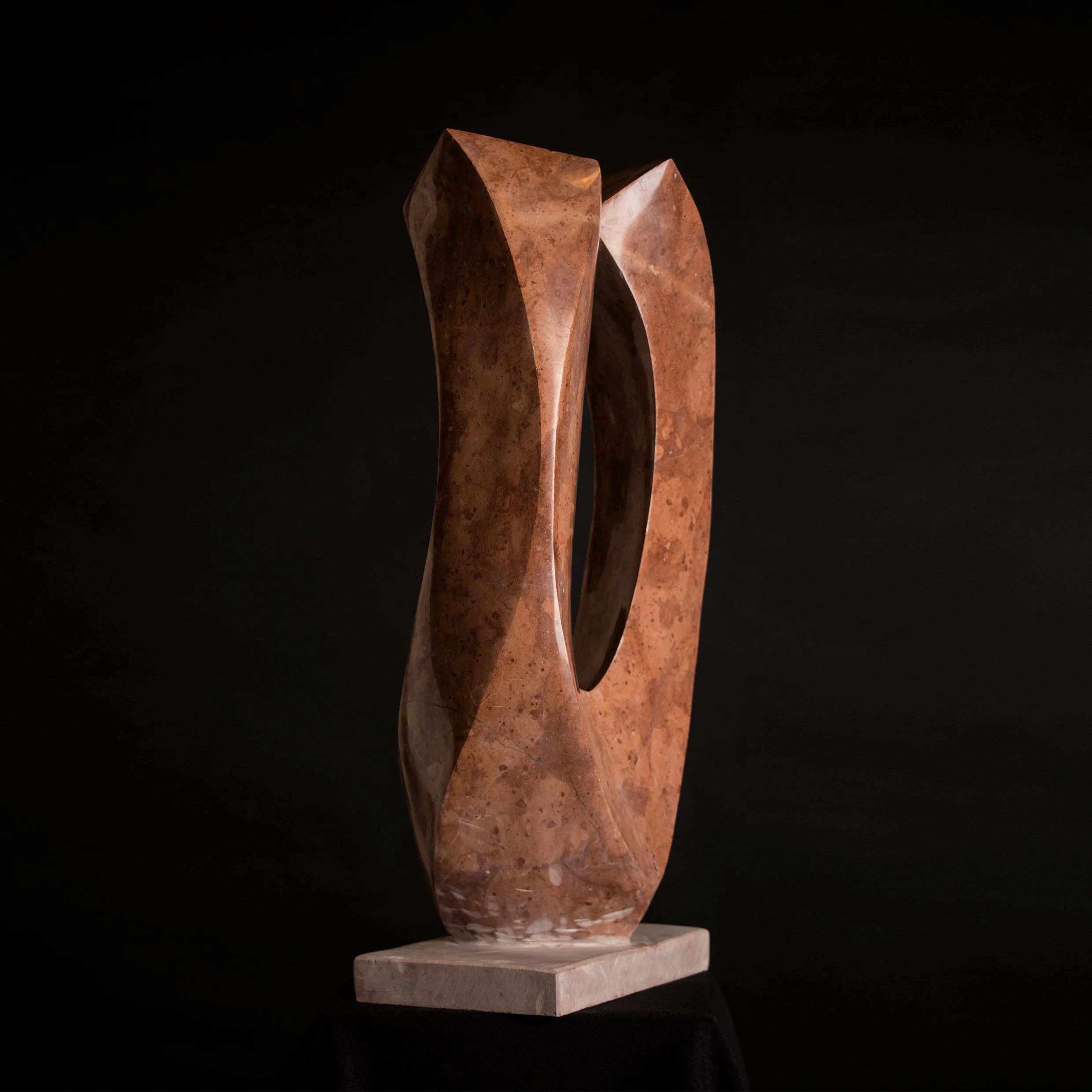 Sculptural artwork made of rust-colored stone with an abstract design, featuring an open oval shape and curved surfaces, mounted on a white rectangular base.