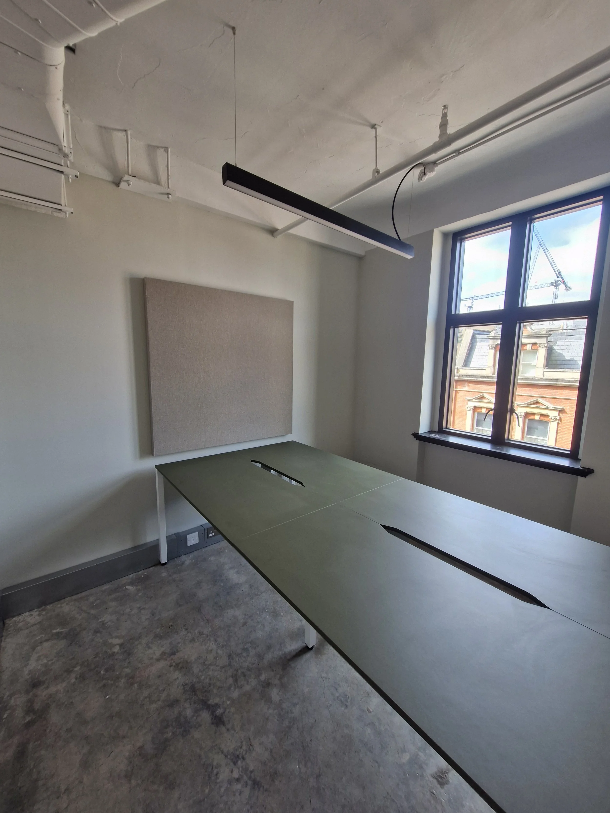 Empty office with a conference table, window with a view of a crane and brick building, and acoustic panel on the wall.