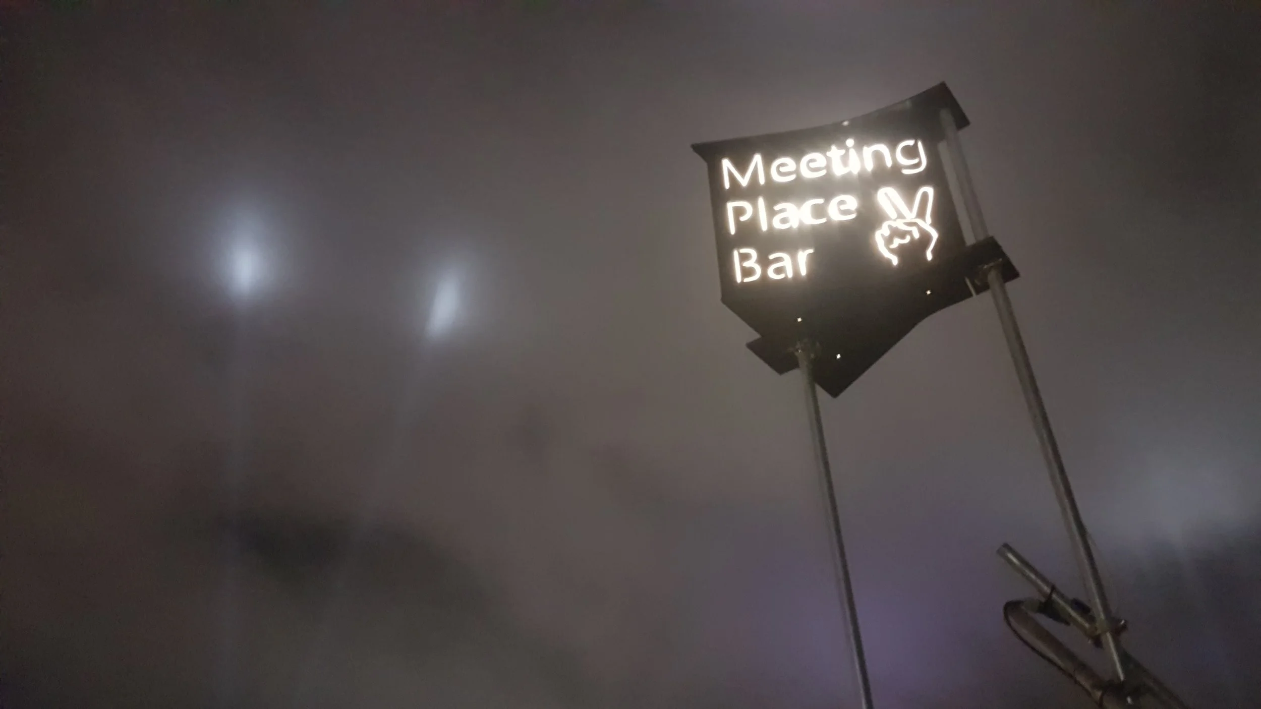 Illuminated neon sign reading 'Meeting Place Bar' with a hand gesture symbol, against a dark cloudy sky at night.