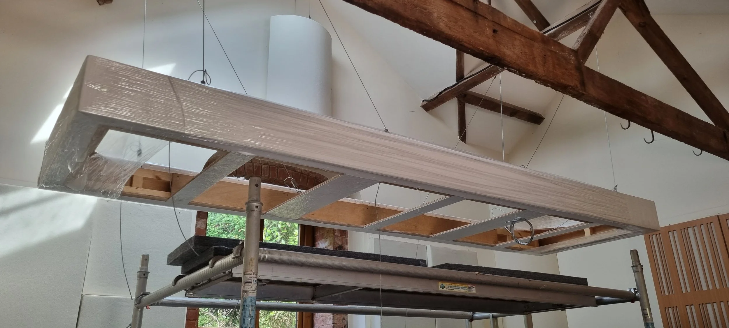 construction site with a suspended wooden framework, metal supports, and work scaffolding inside a room with a window and exposed brick wall.
