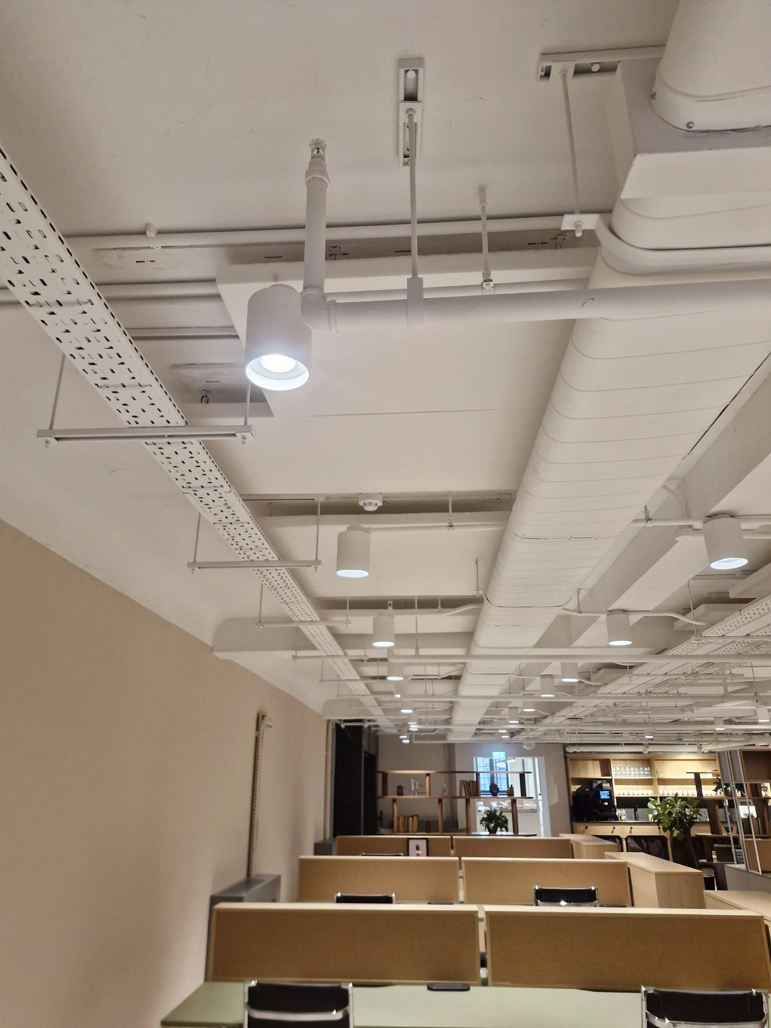 A modern office space with an exposed ceiling featuring white pipes, ducts, and lighting fixtures. There are beige desks and black chairs arranged neatly, with a reception area and shelving visible in the background.