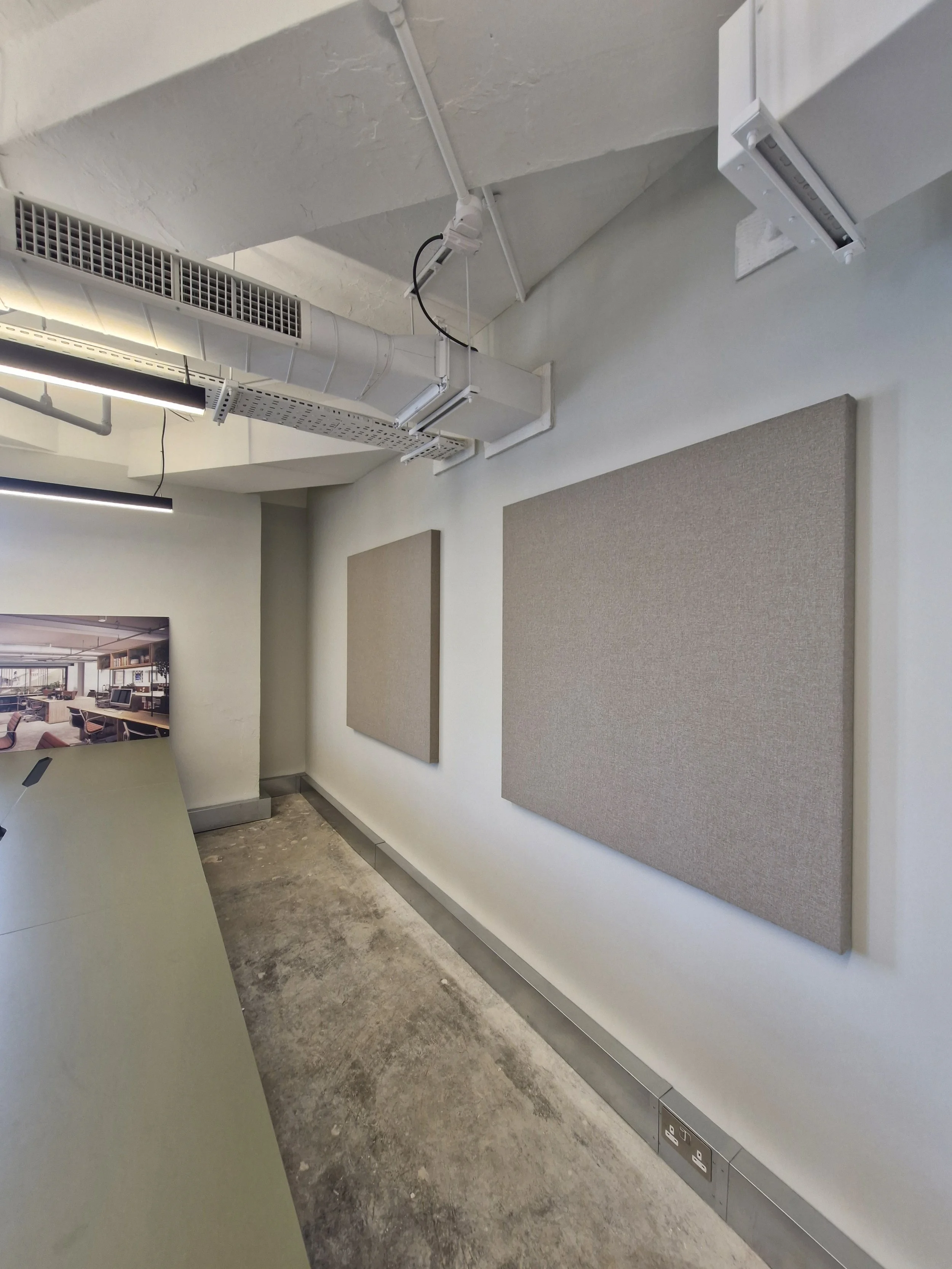 Interior view of a meeting or conference room with two beige acoustic wall panels, overhead industrial air ducts, lighting fixtures, and a power outlet along the baseboard