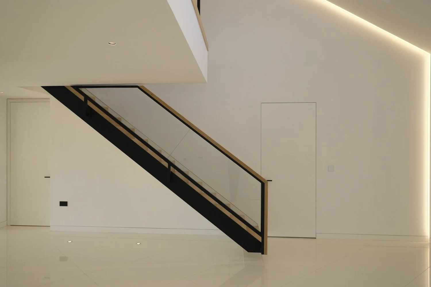 Interior view of a modern, minimalistic white room with a staircase featuring wooden handrails and glass balustrades, illuminated by recessed ceiling lights and LED strip lighting along the ceiling edge.