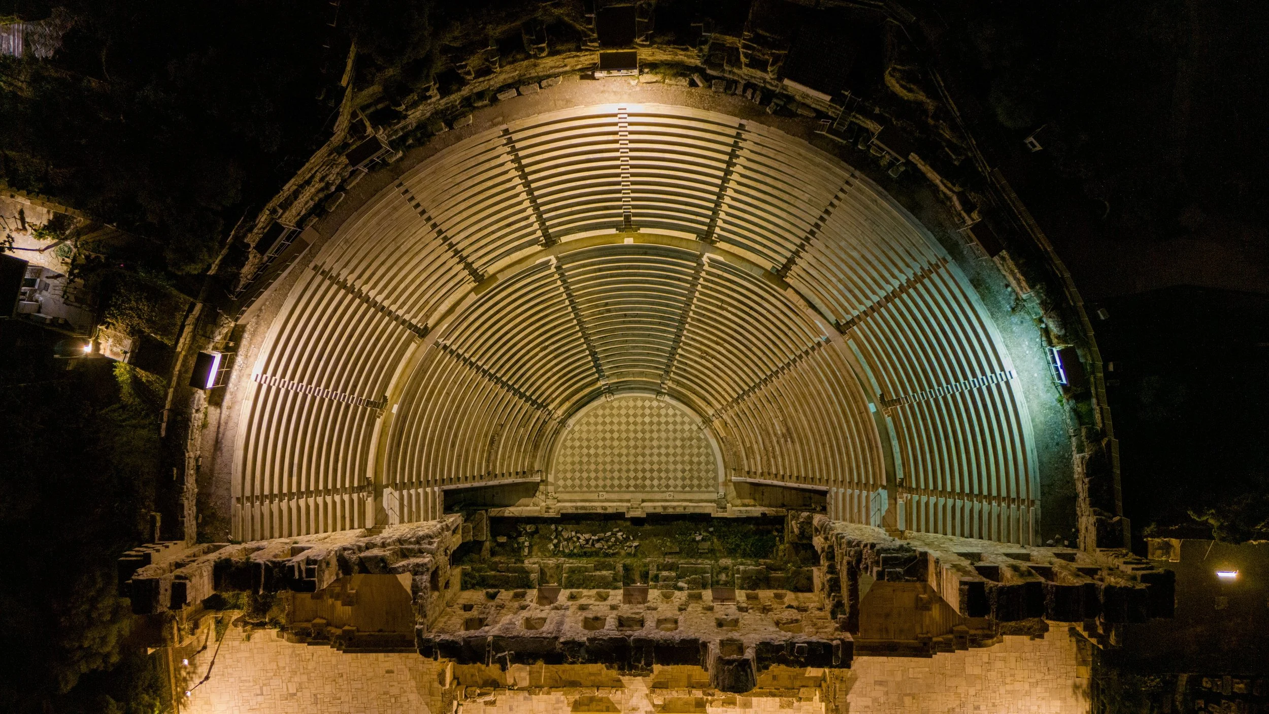 Nighttime aerial view of an ancient, circular stone amphitheater with tiered seating, illuminated by surrounding lights.