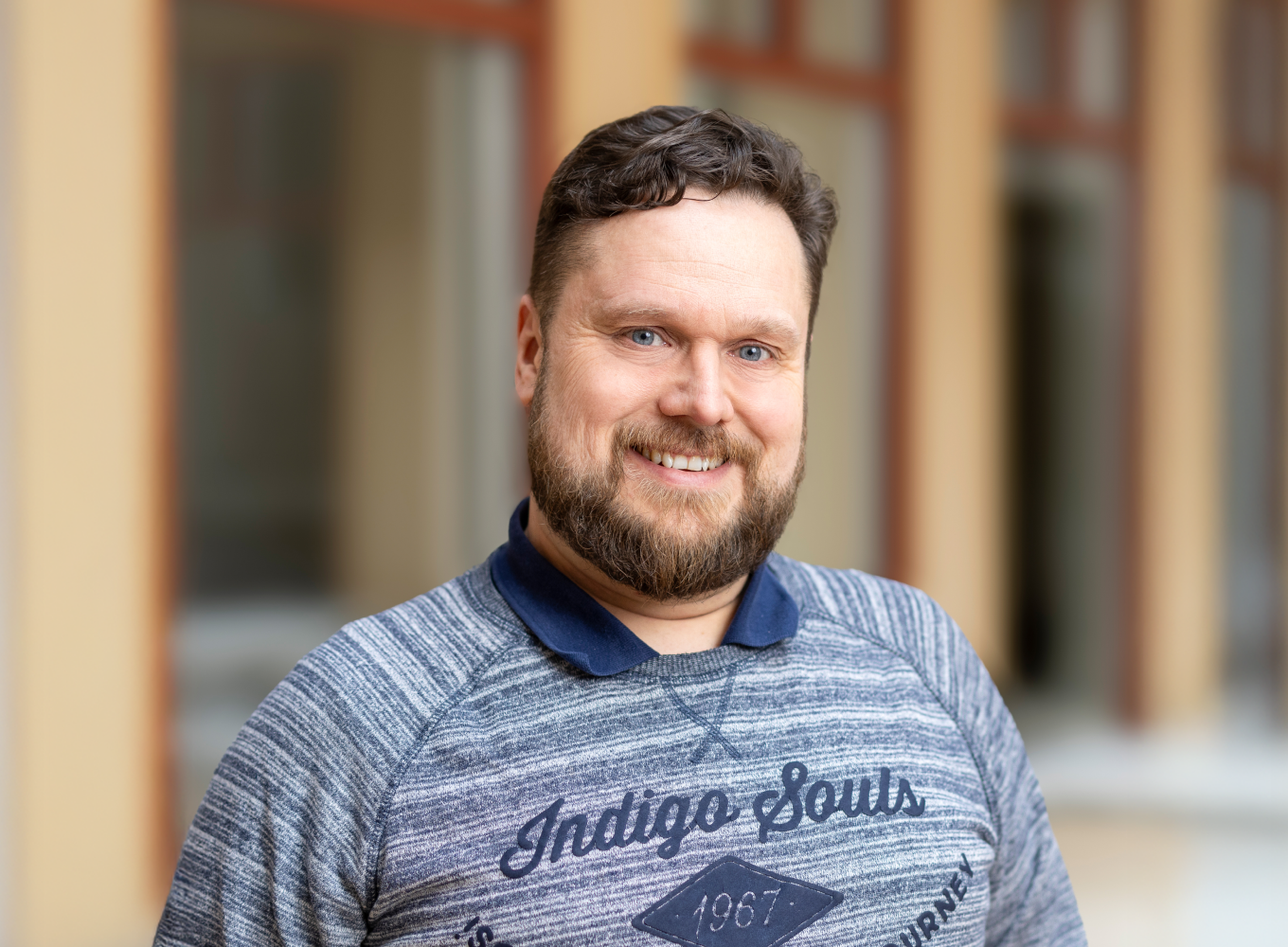 En man med brunt skägg och lockigt hår, ler och tittar mot kameran, står utomhus framför en byggnad med fönster.