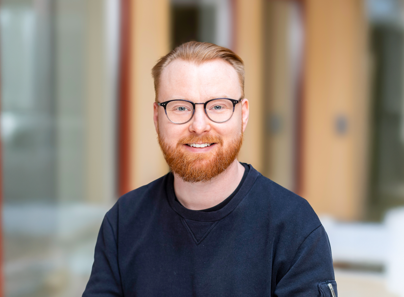 En man med rött hår och skägg, bär glasögon och en svart tröja, står i en inomhusmiljö.