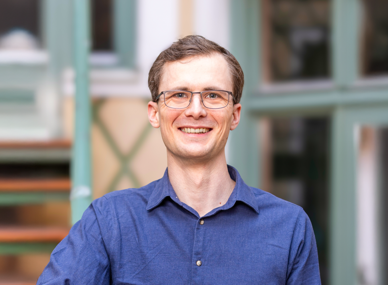 Ung man med glasögon och blå skjorta, är glad och utomhus framför ett trädgårdsbyggnad.