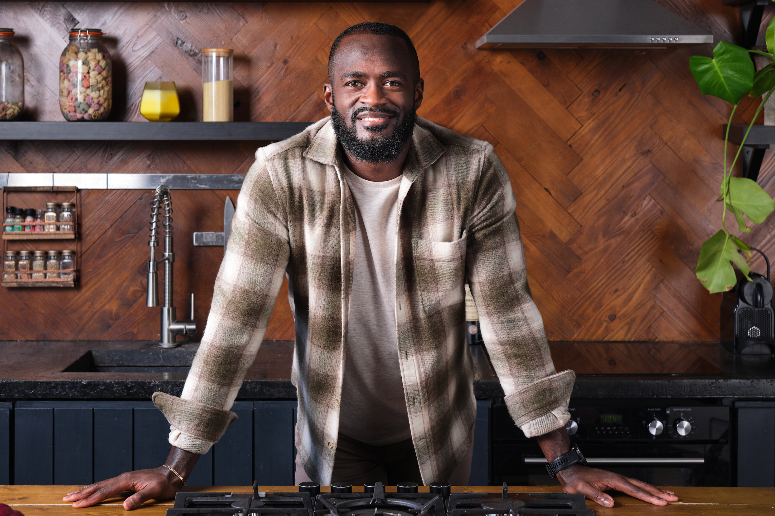 A smiling man in casual clothing leaning on a kitchen counter in a modern kitchen with wooden walls and shelves with jars and containers.