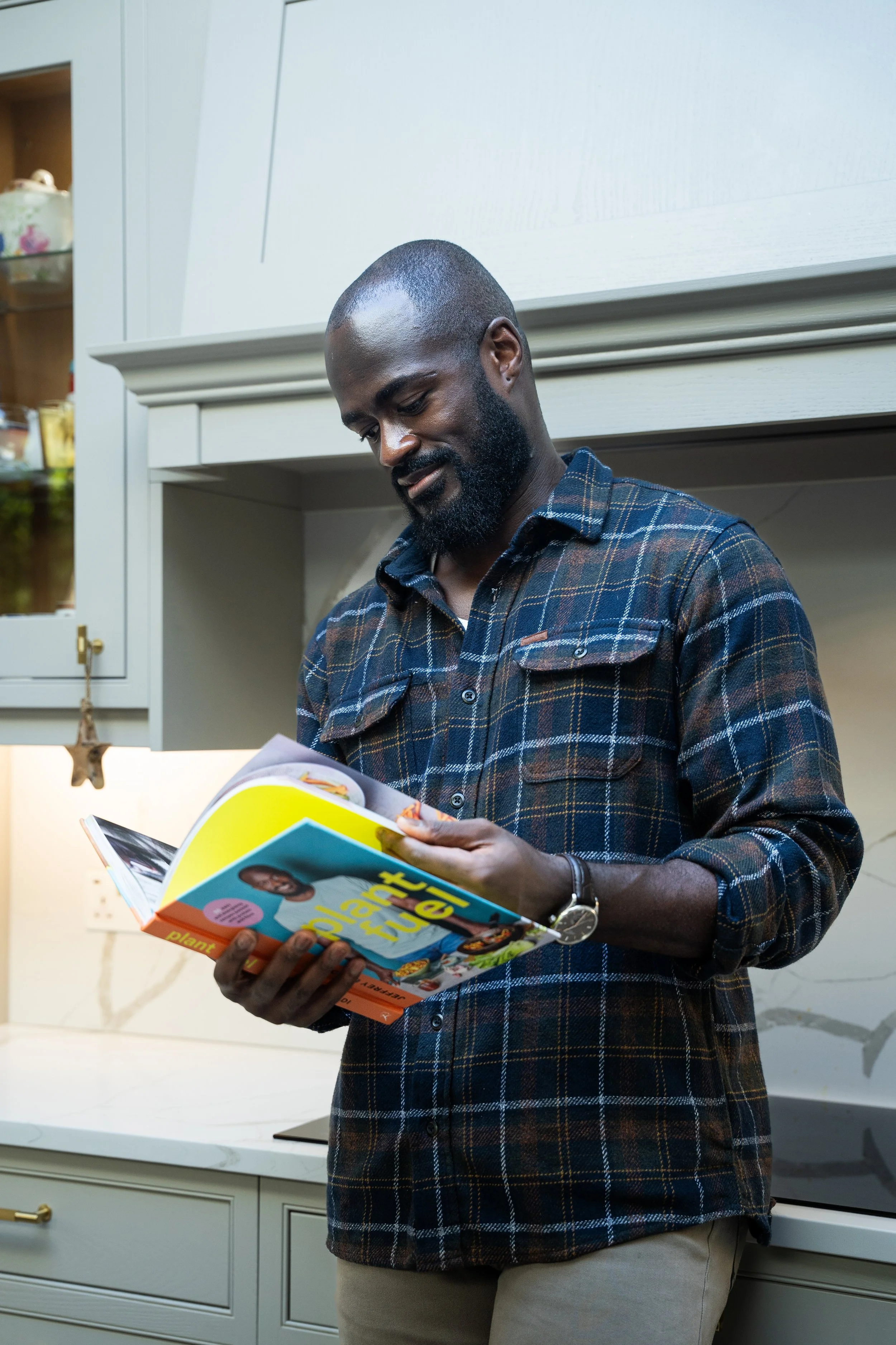 A man with a beard wearing a plaid shirt reading a colorful magazine in a kitchen.