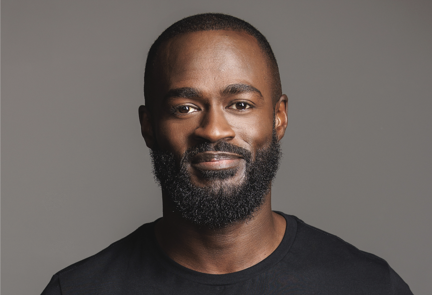 Portrait of a smiling Black man with a beard, wearing a black shirt, against a gray background.