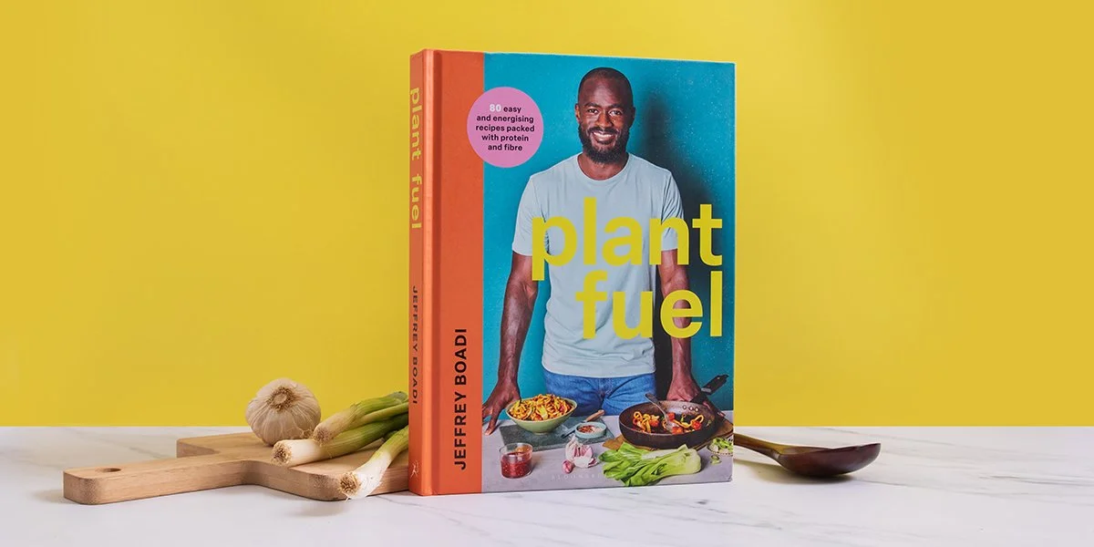 A book titled "Plant Fuel" by Jeffrey Boadi stands upright on a white surface, with fresh garlic, leeks, and a wooden cutting board on the left and a wooden spoon on the right. The book cover features an image of a smiling man in a white t-shirt standing behind a variety of plant-based dishes against a teal background, with a yellow wall in the background.