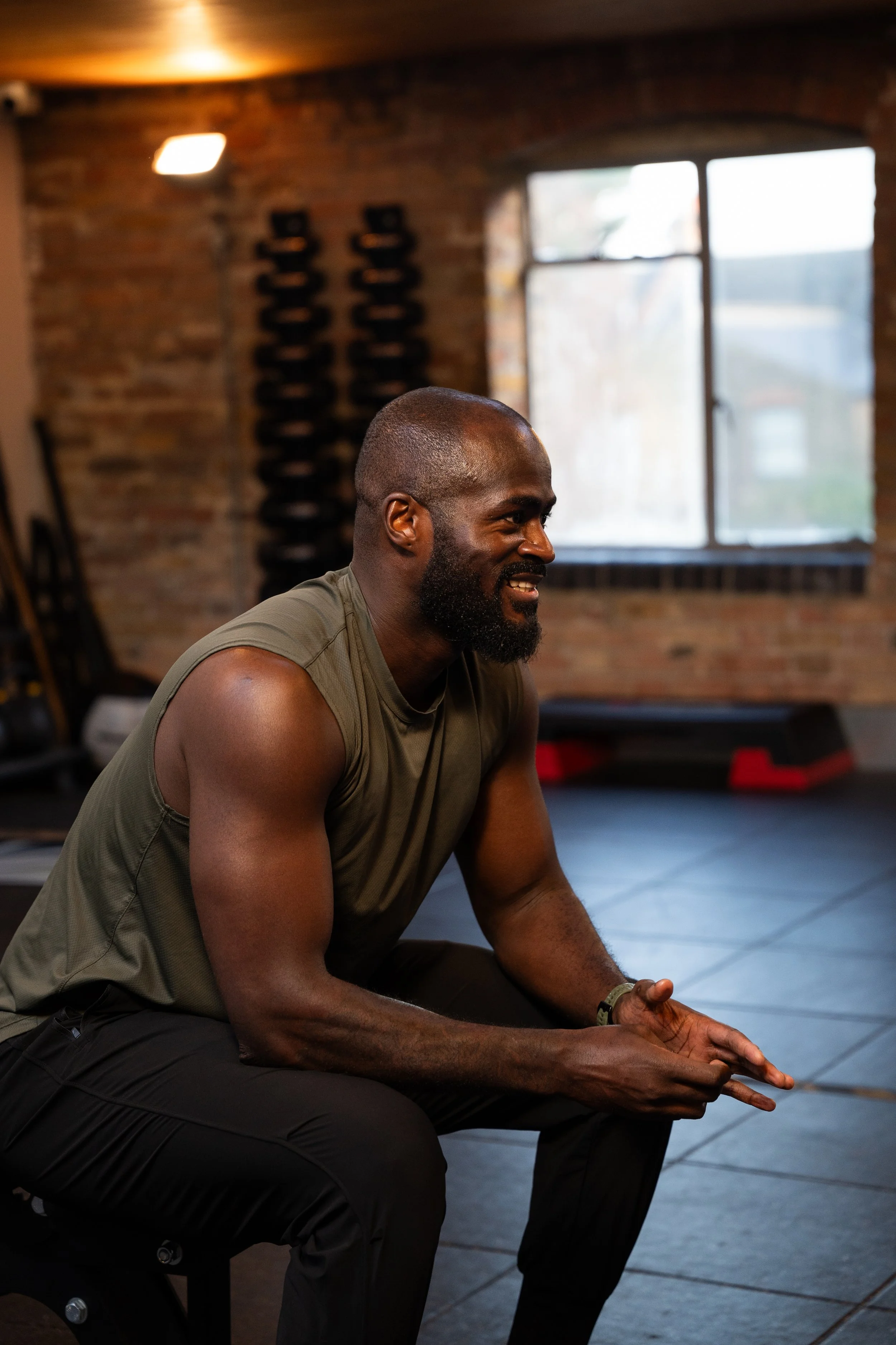 A man with a beard and bald head, wearing a sleeveless shirt, sitting in a gym with brick walls, black equipment, and a window in the background.