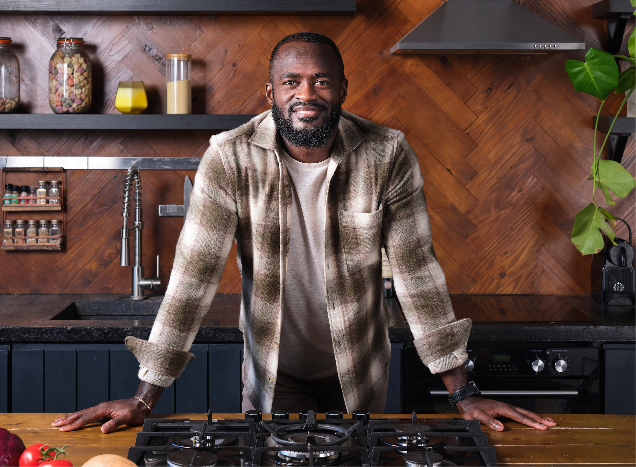 A man in a flannel shirt leaning on a kitchen island, smiling, in a modern kitchen with wood-paneled walls and various jars and kitchen utensils.