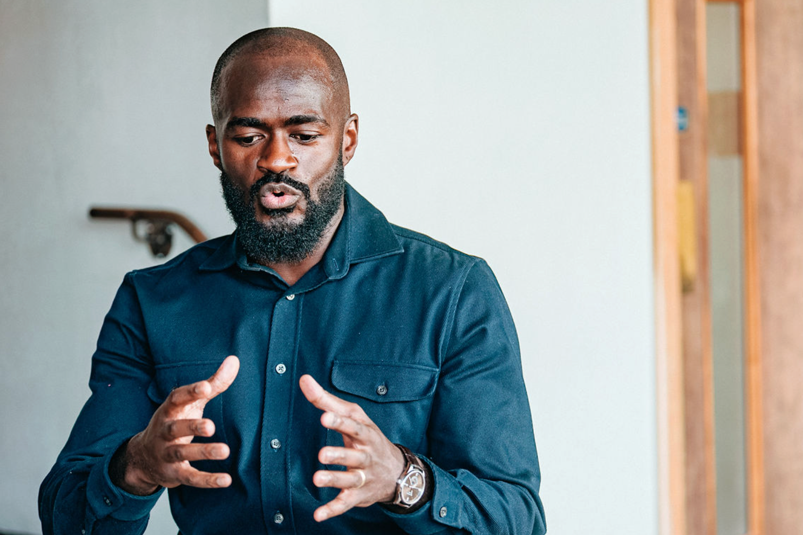 A man with a beard and short hair wearing a dark blue button-up shirt gestures with his hands while speaking.