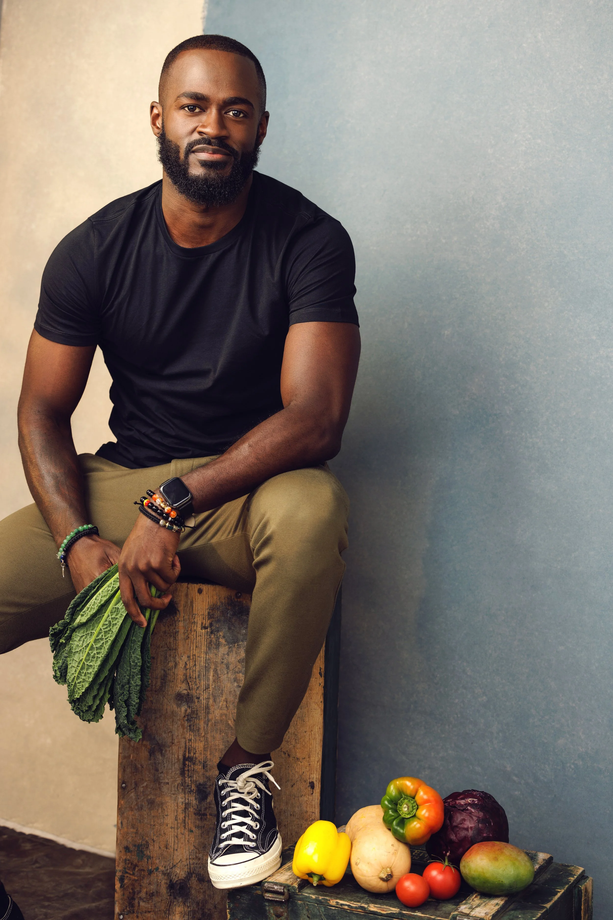 A man with a beard and short hair, wearing a black T-shirt, khaki pants, and black-and-white sneakers, sitting on a wooden box, holding leafy greens in his left hand, with various vegetables on a small wooden table nearby.