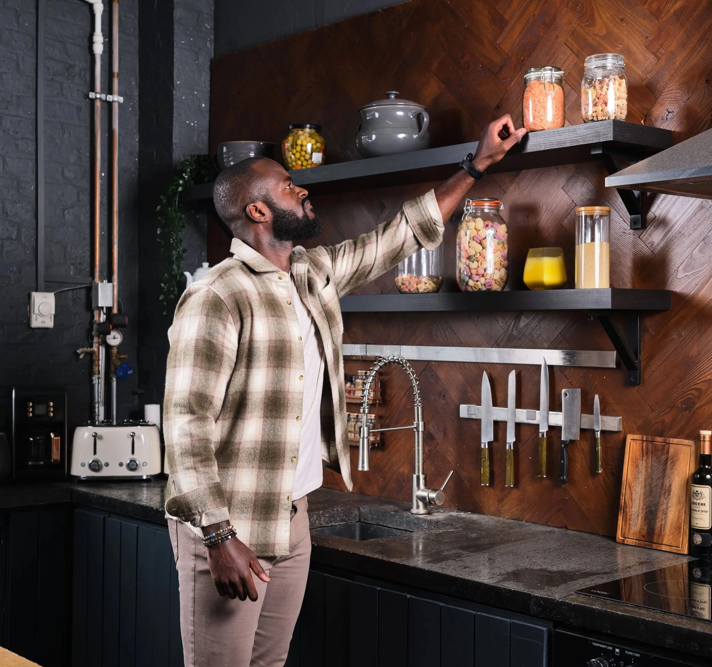 A man reaching for a jar on a kitchen shelf with other jars and kitchen items, in a modern kitchen with dark cabinets and wooden wall panels.