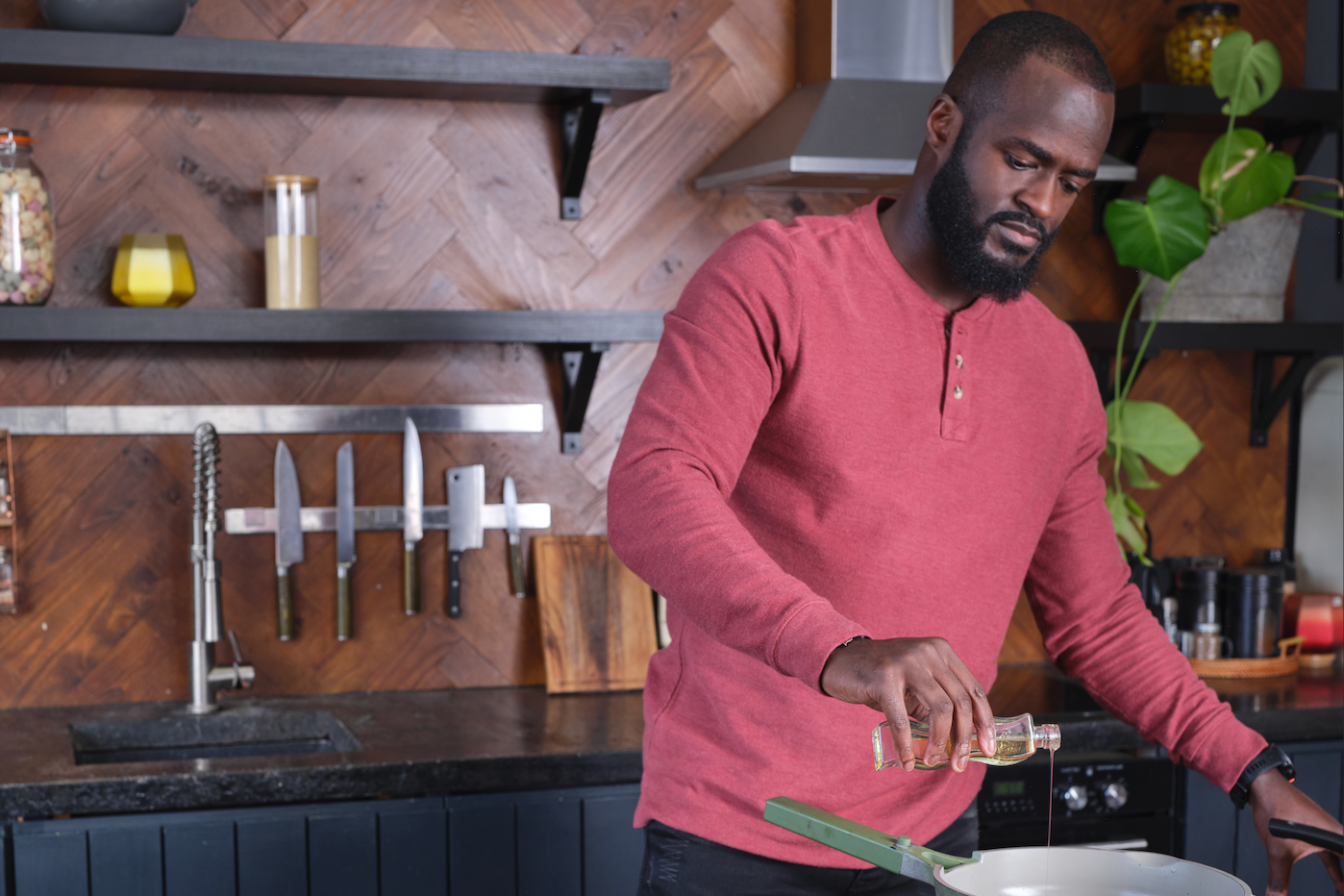 A man in a red long-sleeve shirt adds oil to a pan in a modern kitchen with wooden walls and shelves, knives on a rack, and a plant in the background.